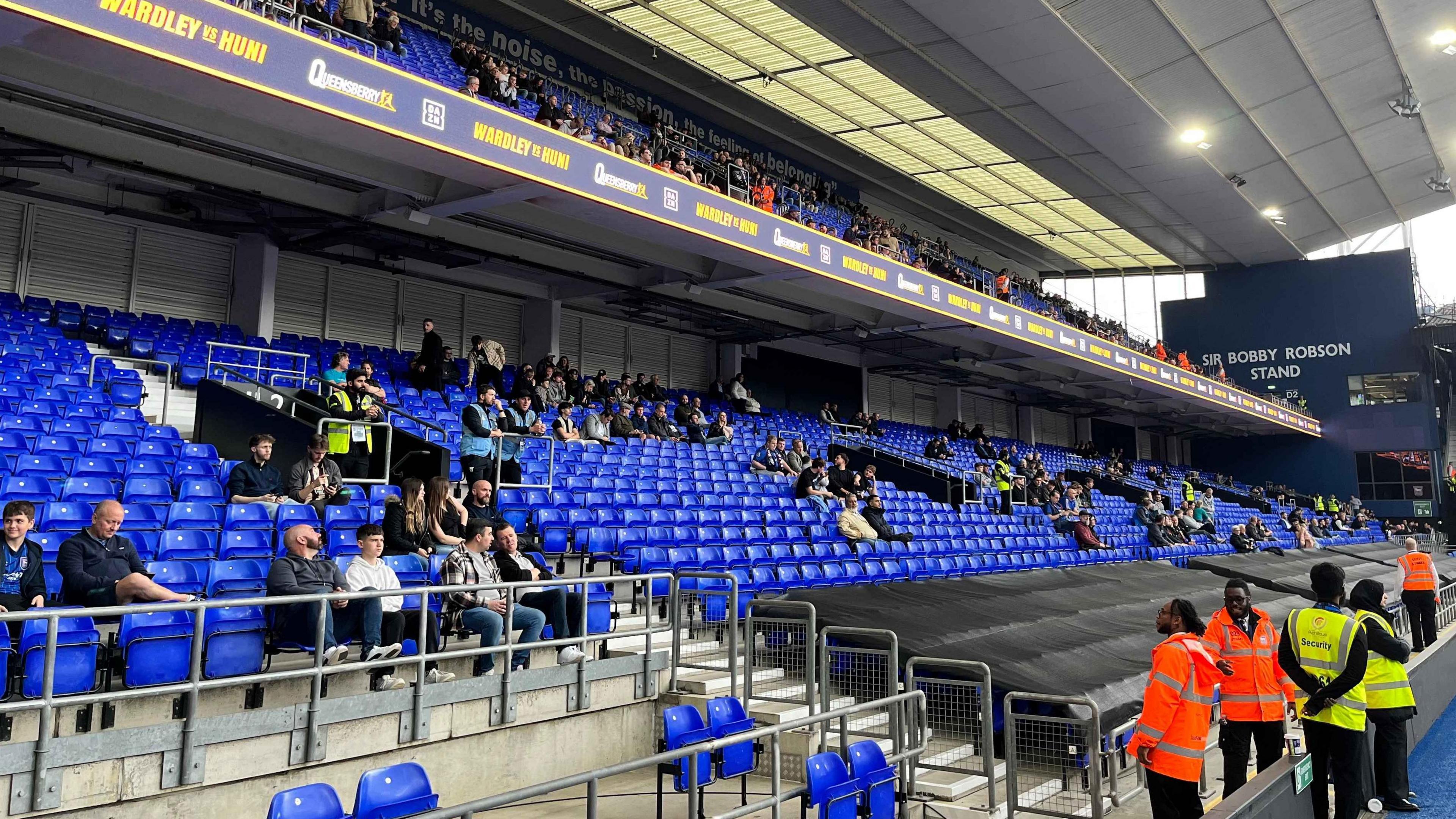 A close up view of one of the stands at the stadium that has two levels. The chairs are blue, and in long rows which span across the whole picture. People are sat staring towards the right of the image where the boxing ring is.