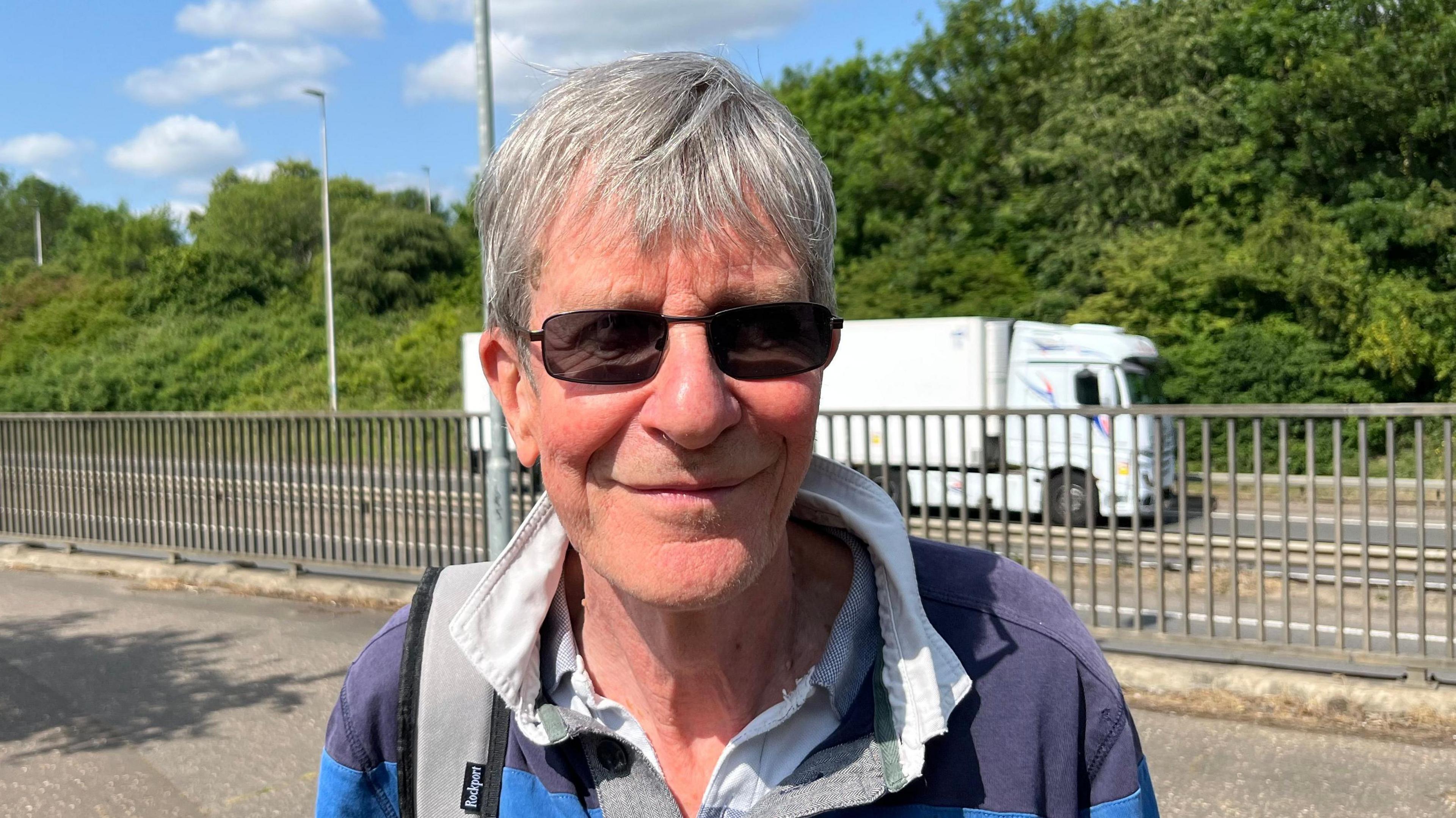 Harry Machin has short grey hair swept across his forehead. He has dark glasses on and a light blue and dark blue hooped rugby style shirt on, with a white shirt underneath. Behind him are metal railings and a busy roadway, with a white lorry passing.