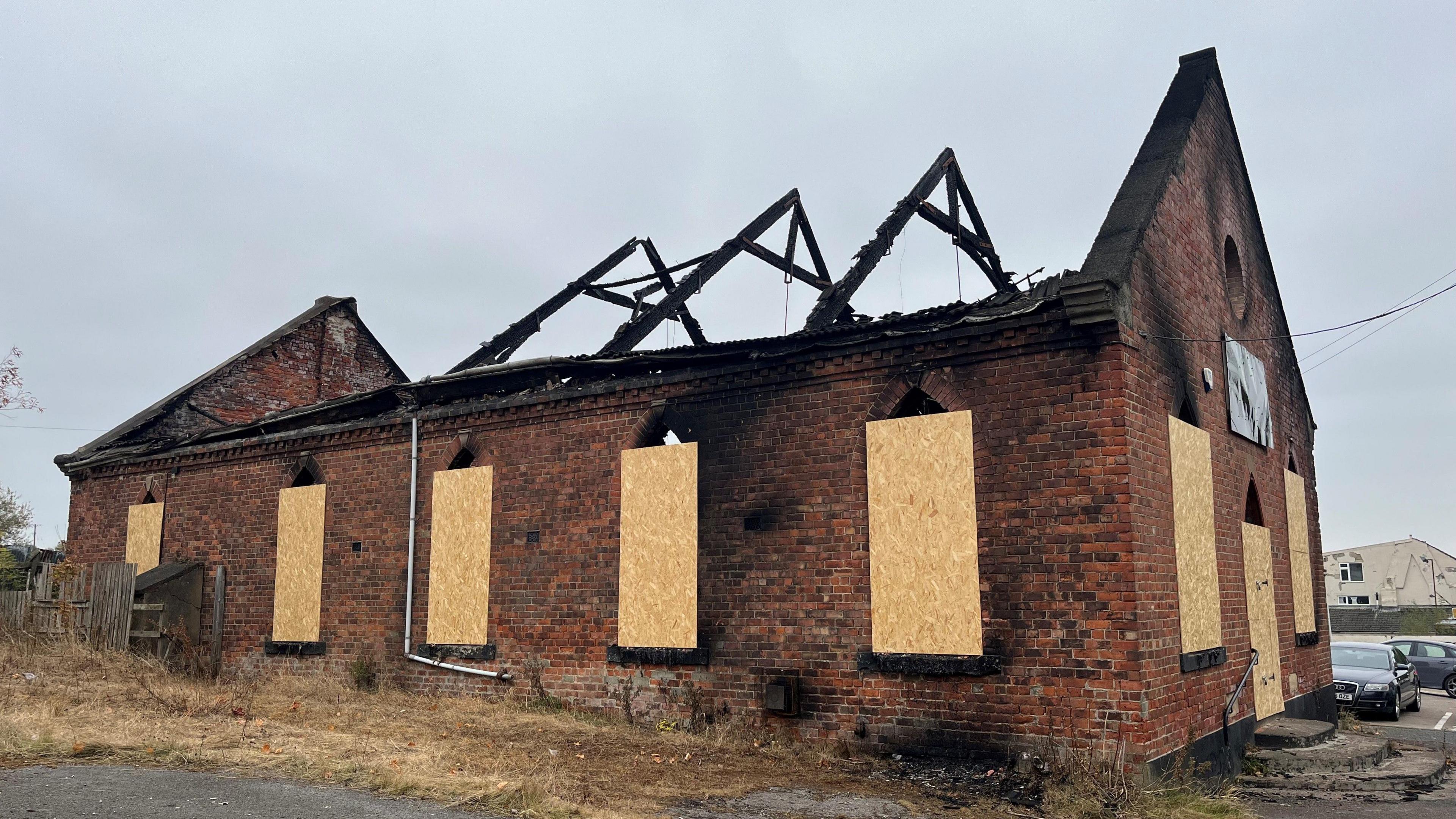 The exterior of the burnt-out brick building, the windows are boarded up and burnt wooden beams exposed.