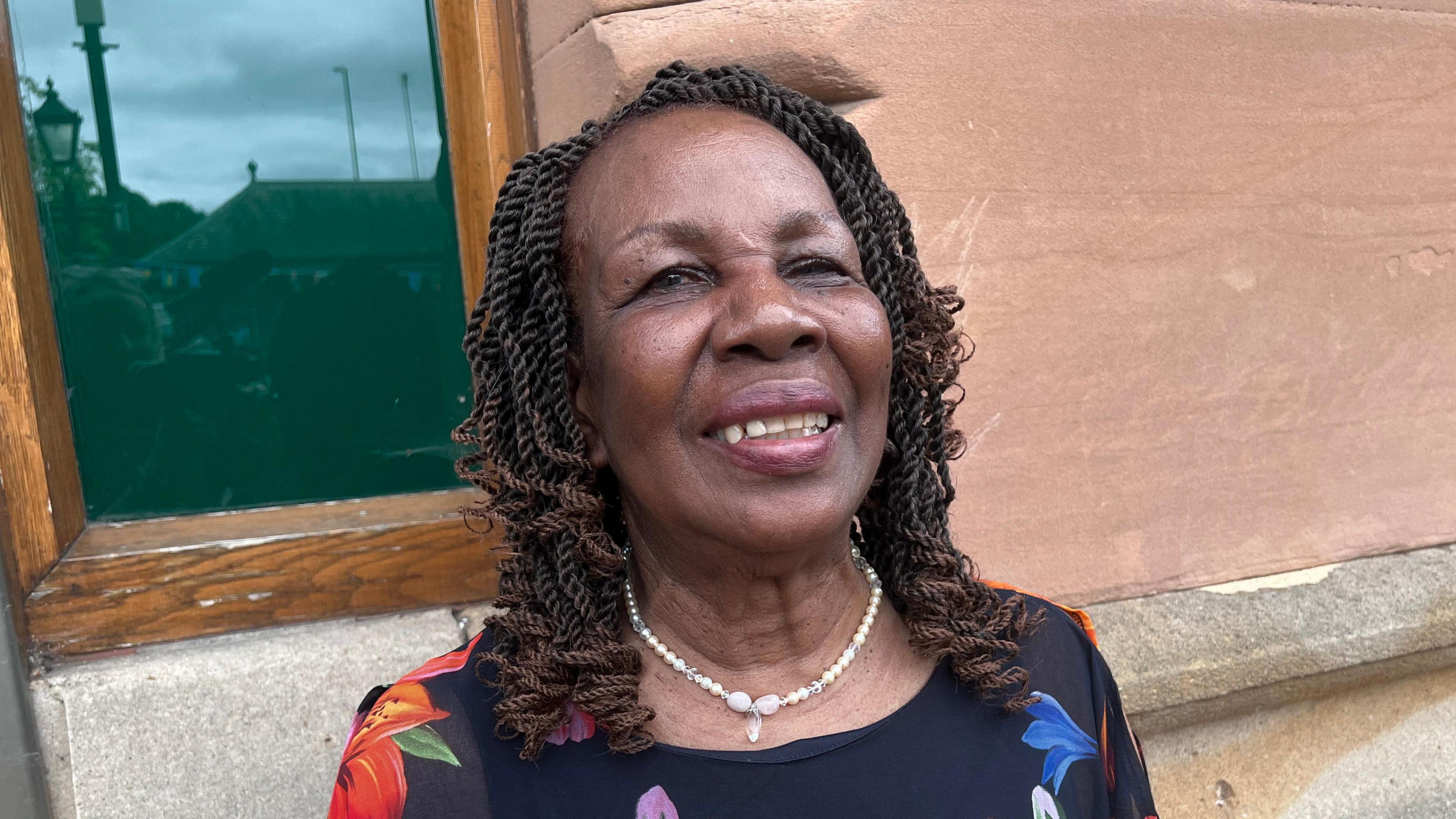 Lilian Francis is standing in the middle of the picture looking slightly to the left of the camera. She is wearing a black top with floral patterns on and has a necklace made of white pearls.