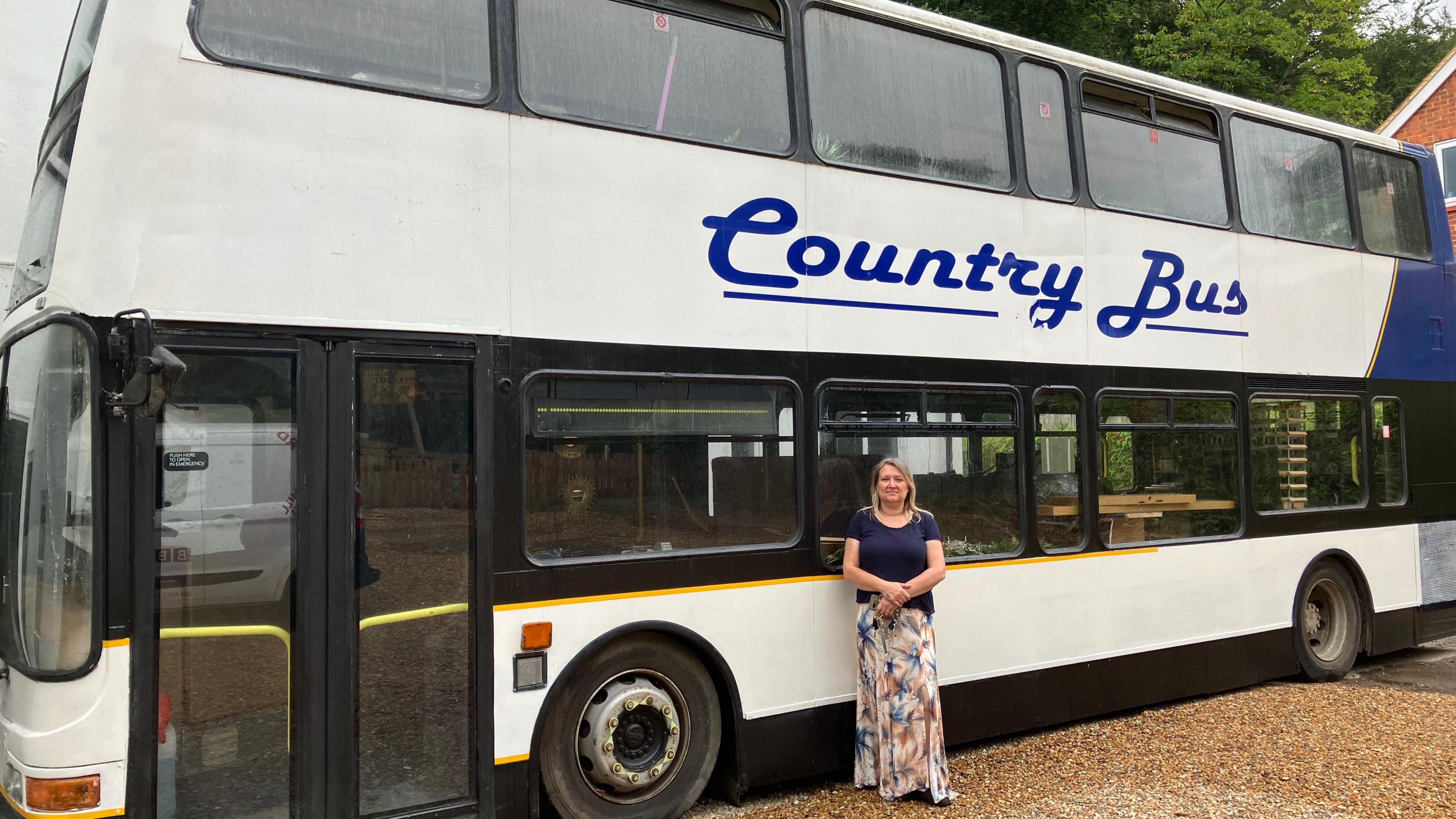 Wendy Salmon stands outside the double decker bus, she is wearing a dark blue t-shirt and a long patterned skirt. The bus is mostly white, with a blue streak down the back and the words Country Bus on the side in blue lettering.