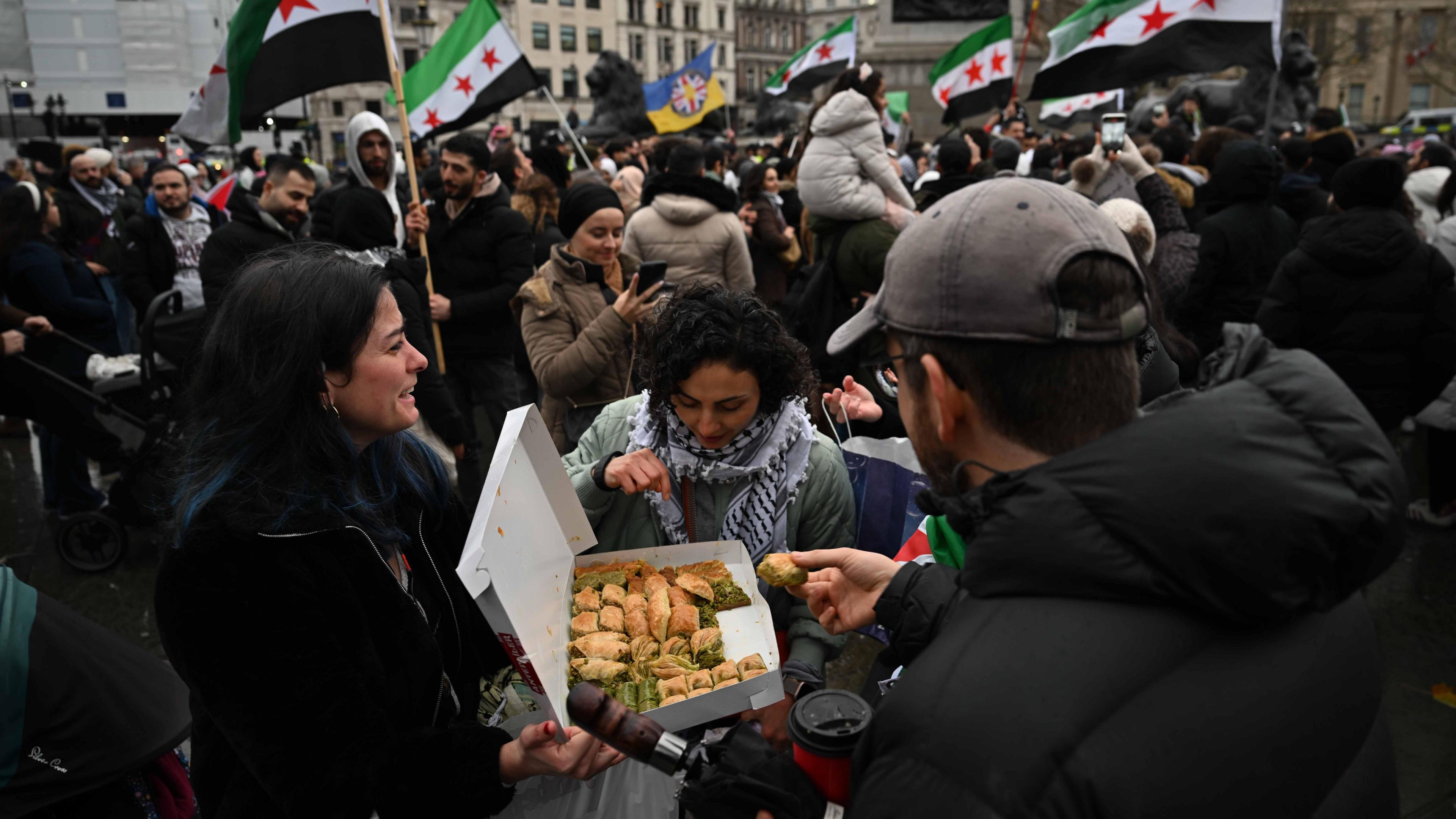 An image of the demonstration at Trafalgar Square where people hold Syrian flags in the background. A woman hands out baklava and another woman and man reach in to take a bite.