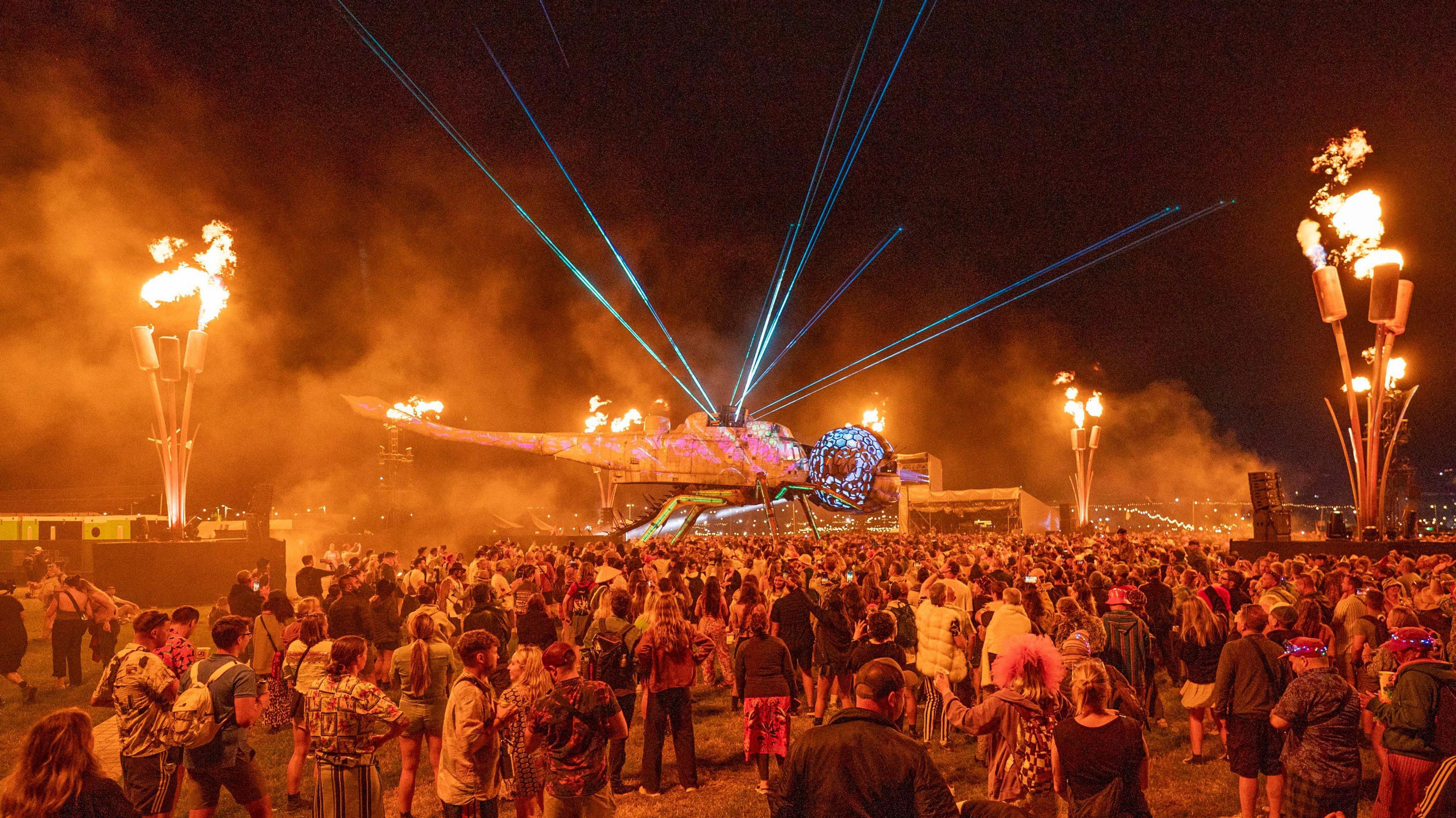 A crowd gathers underneath a dragonfly which has a helicopter as a body. Lasers shine from the dragonfly's body and there are flames burning around the edge of the crowd. The flames are so strong, they are leaving an orange glow above the crowd.