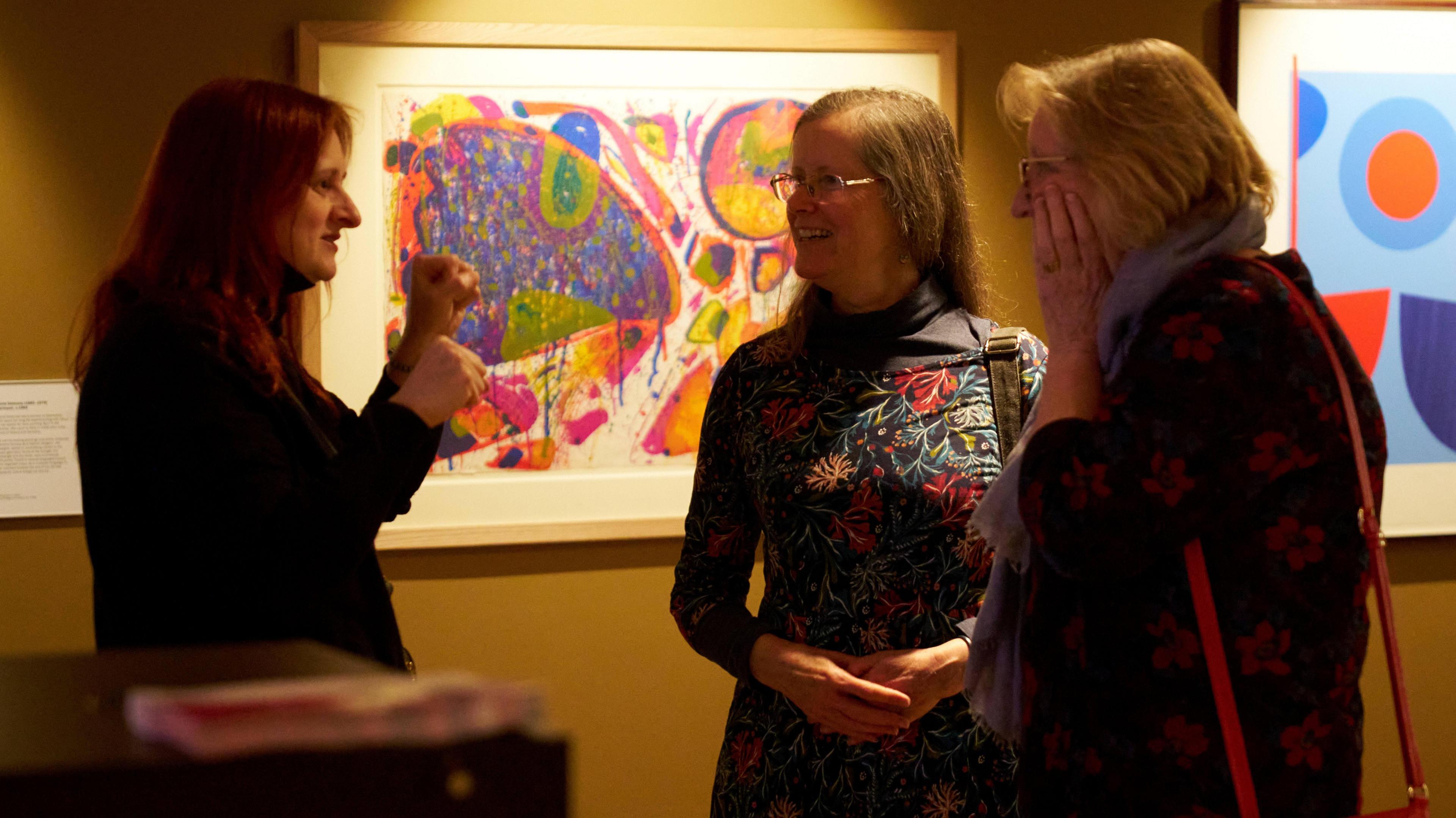 Alison Goodyear (left) talking to Professor Christiana Payne (middle) and Dr Mary O'Neill (right). They are standing in a gallery looking at paintings, Alison is holding up her arms, Chistiana has her arms folded in her middle and Mary has her arm to her face, there is a colourful painting on the wall behind him. 