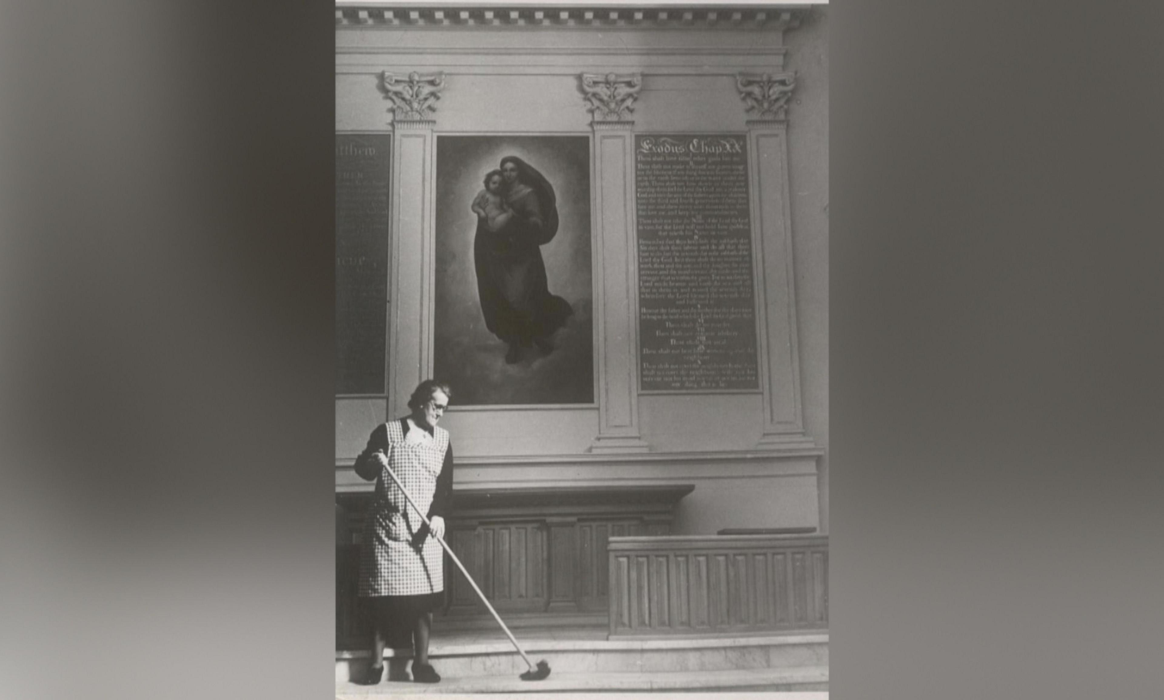 Stood on the steps by the alter, Mabel Wulff is pictured in a black and white photograph sweeping the church floor with a large broom. Dressed in a cheque pinafore over a black dress, the post-war photograph depicts her efforts to bring the church back to life after months of battle in the city of Hamburg.