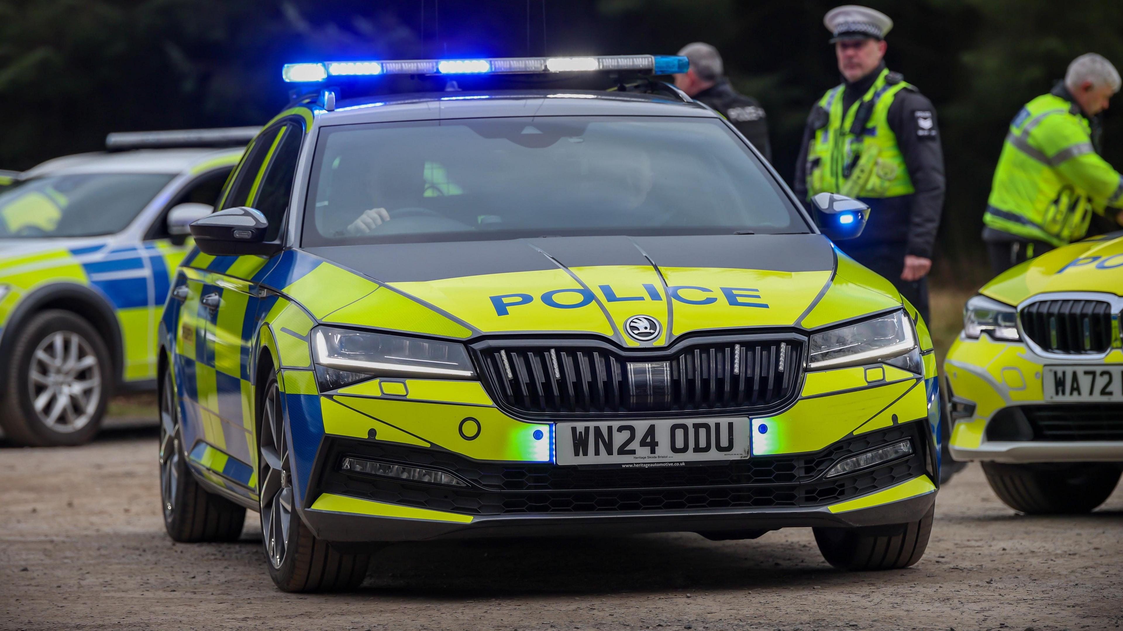 A black, yellow and blue Skoda police car from Devon and Cornwall Police leaves a car park with its blue lights on. Its licence plate is WN24 0DU. Two officers are sat inside the car. Two other police cars are parked up nearby. Three officers - two of whom are wearing high-vis jackets - are stood next to the other cars.