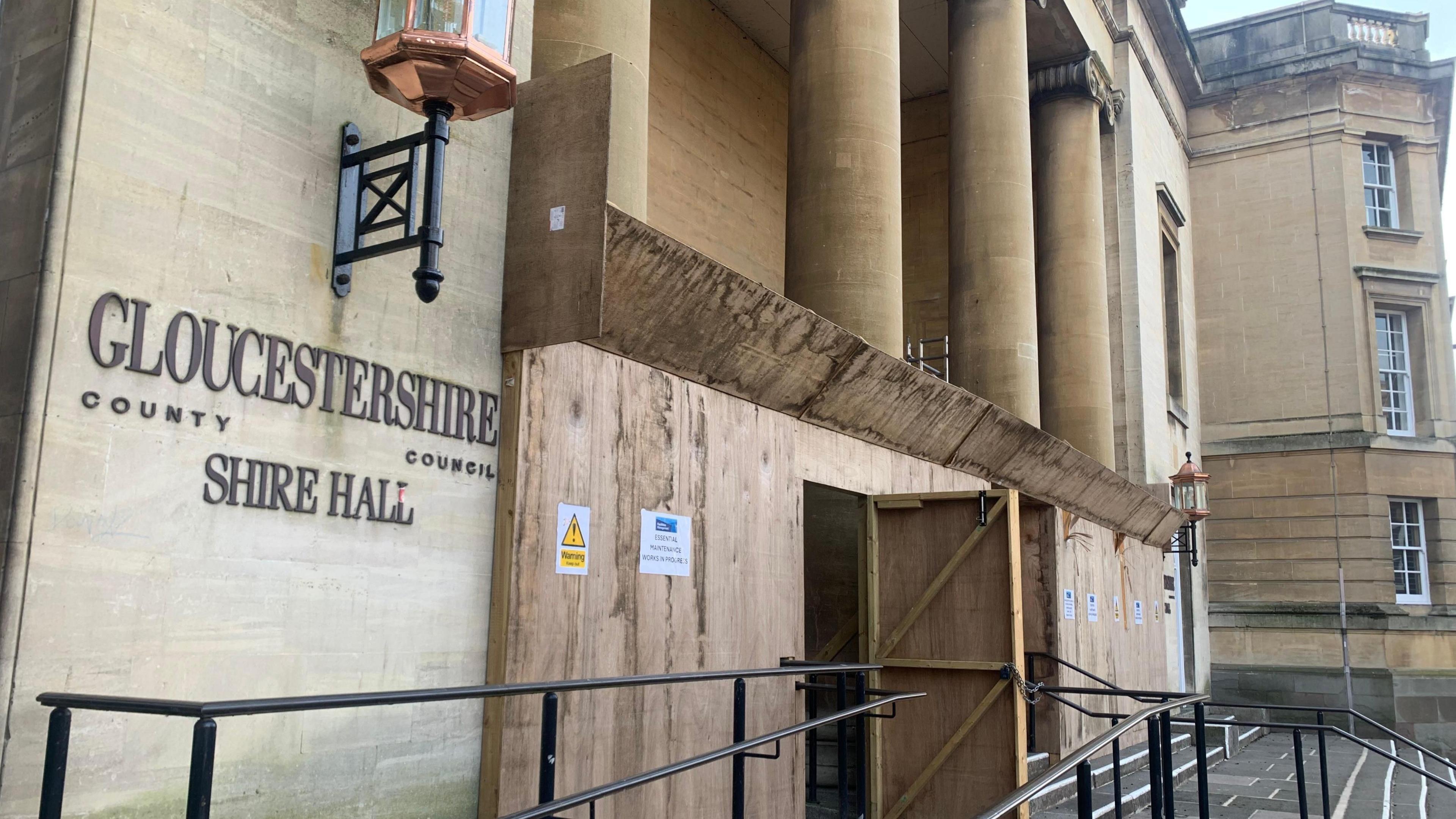 An exterior view of Shire Hall in Gloucestershire. The building is covered in scaffolding, and is approached by a grey stone staircase. The sign on the building reads 'Gloucestershire County Council Shire Hall'. 