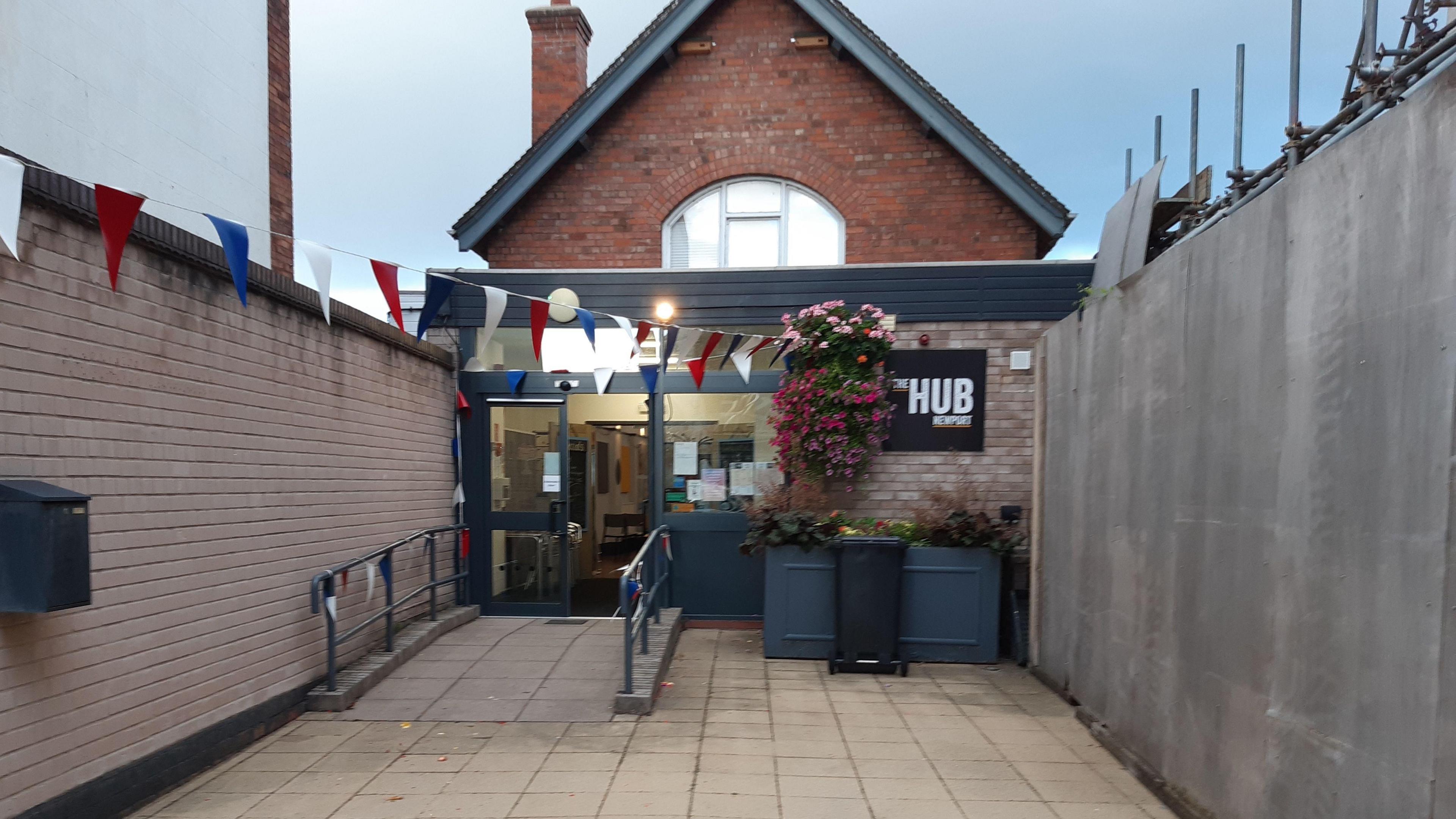 A brick building with a black sign reading "The Hub Newport" and flowers in baskets and planters outside. It has glass doors and walls either side of it, and railings immediately outside.