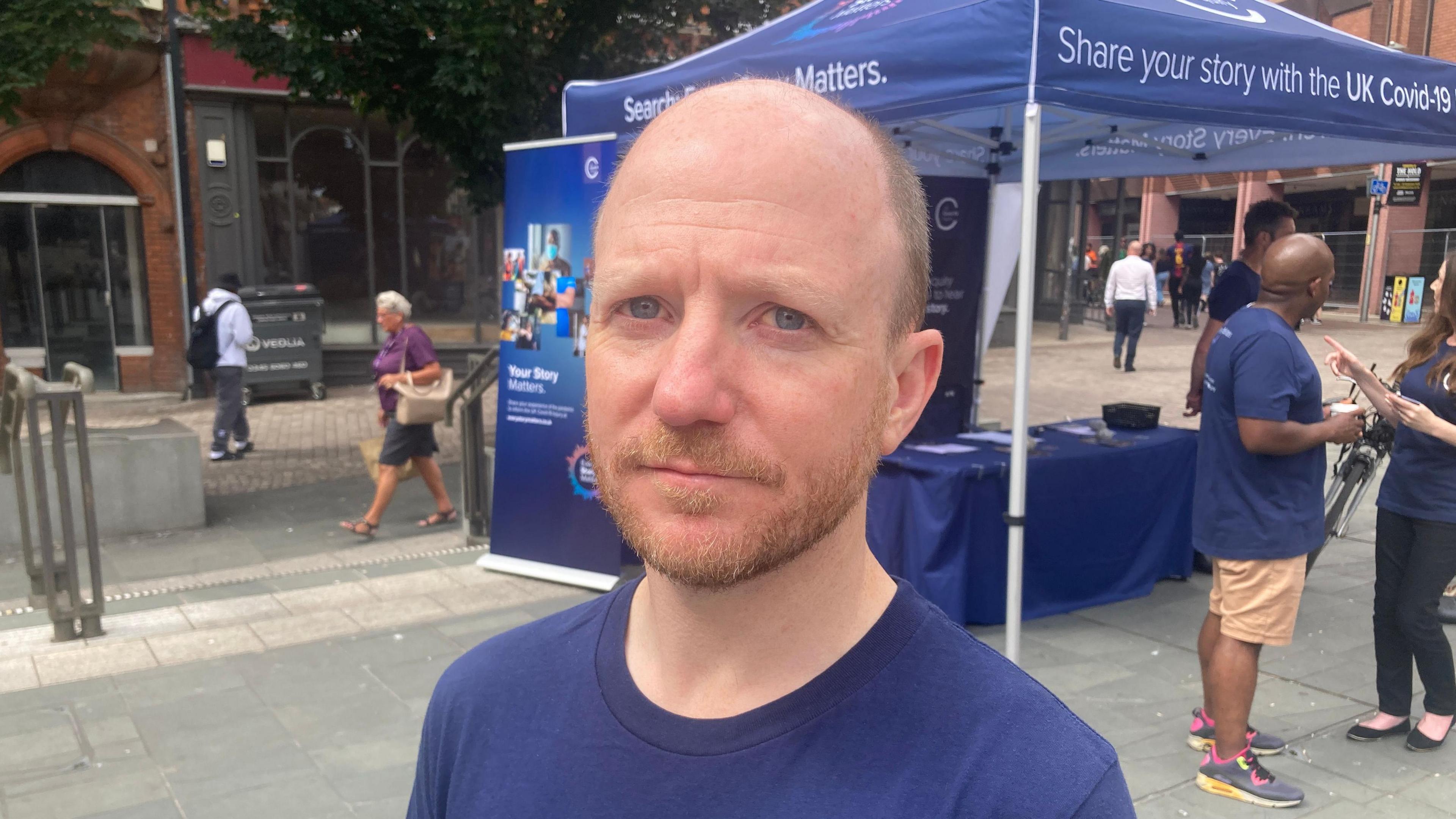 Ben Connah, a bald man with a beard who is wearing a blue t-shirt, stands in front of an inquiry-branded tent where people can give their pandemic stories to inquiry staff