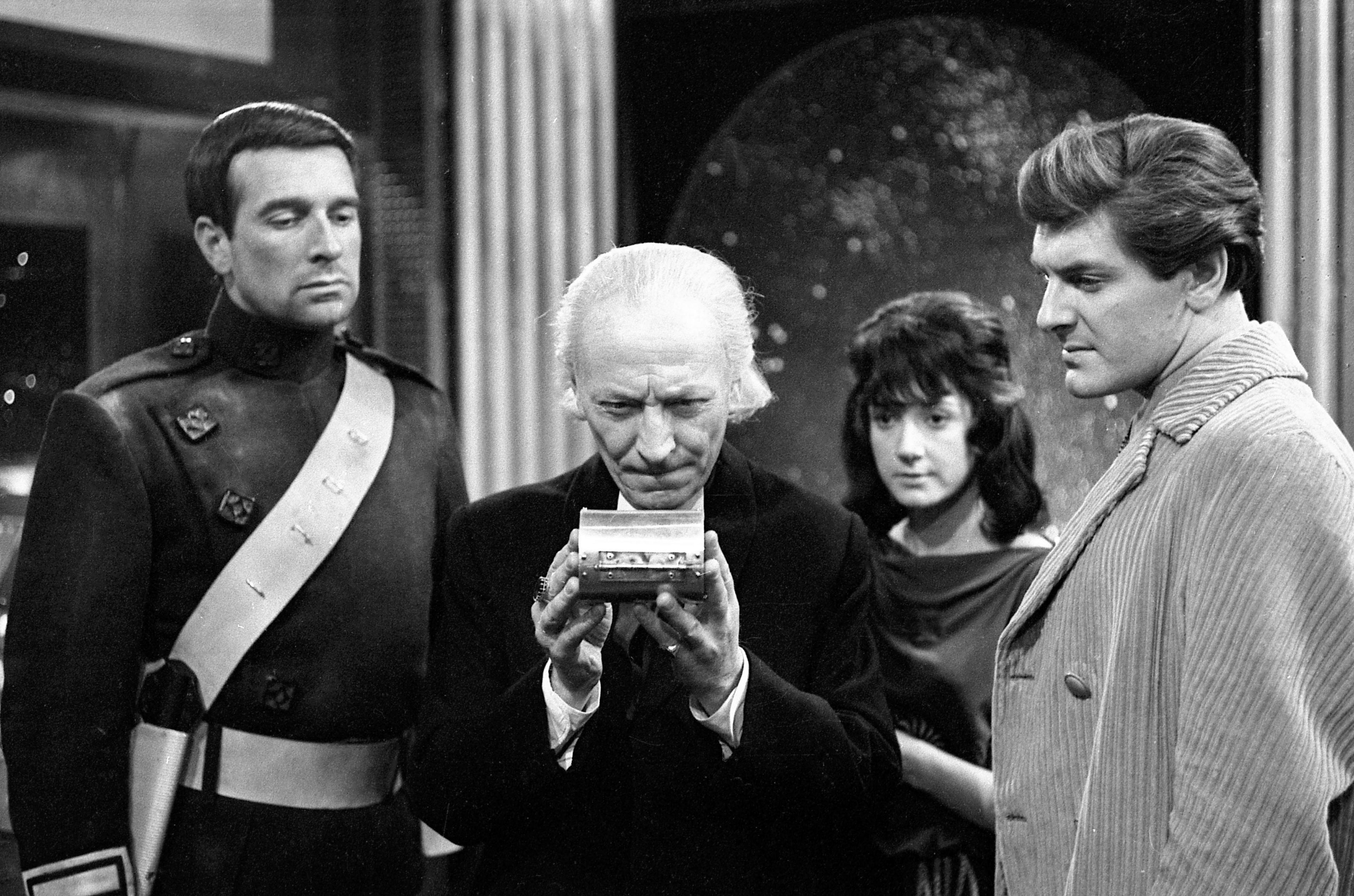 Black and white photo from the set of The Daleks' Master Plan. William Hartnell, with white hair and a dark coat, as the Doctor is in the centre of the group. To his right is Nicholas Courtney as Bret Vyon, who has short dark hair and is wearing a military style uniform. To the Doctor's left is Peter Purves as Steven, who has dark hair and is wearing a light coloured corduroy jacket. Behind the Doctor's left shoulder is Adrienne Hill as Katriana, who has long dark hair and wears a dark ancient greek style dress. The Doctor is holding up a small metal object which he and those surrounding him are staring at intently.