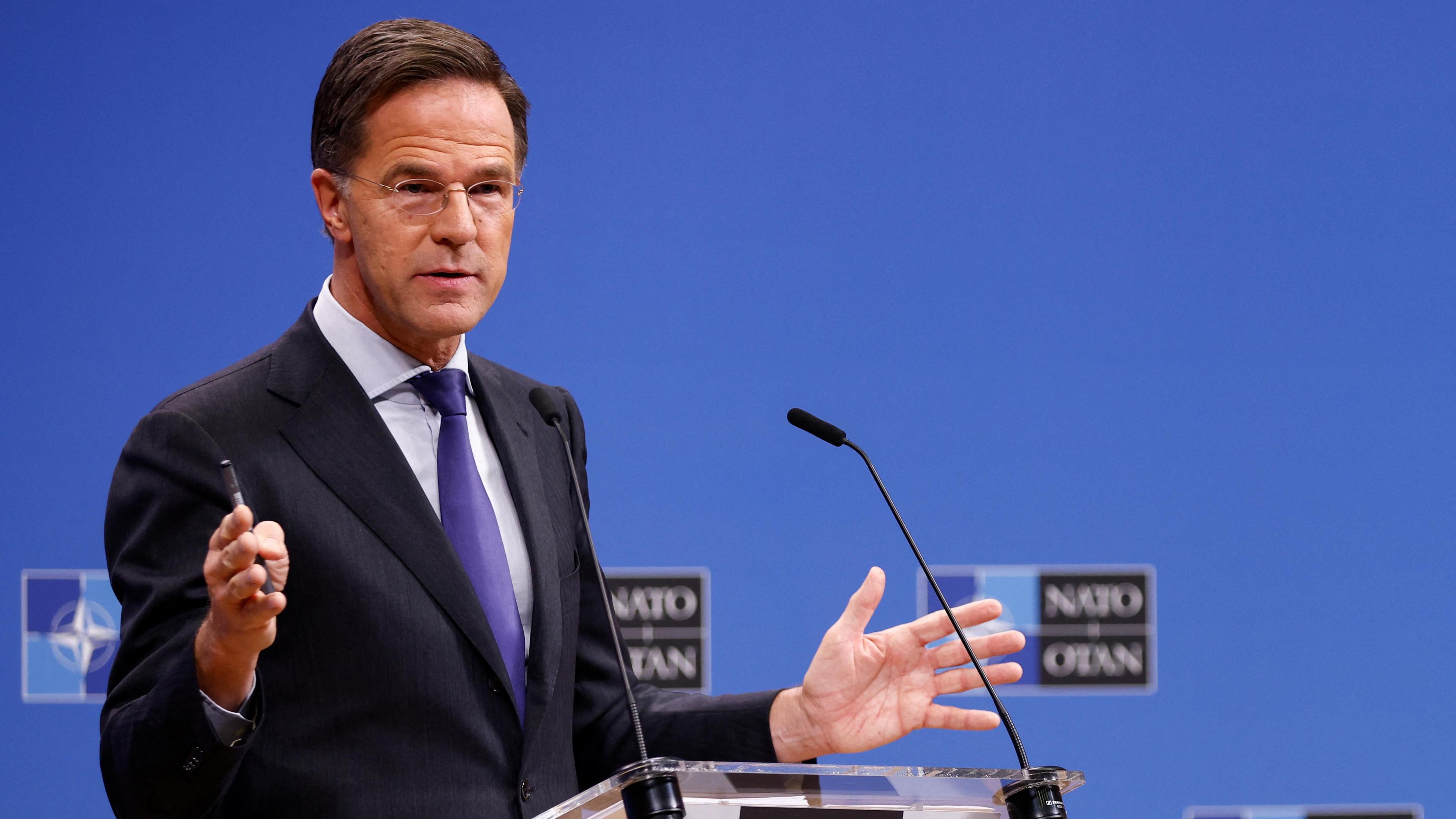 Mark Rutte wears glasses, a black suit jacket and a purple tie. He speaks into a small microphone on a podium in front of a blue background.