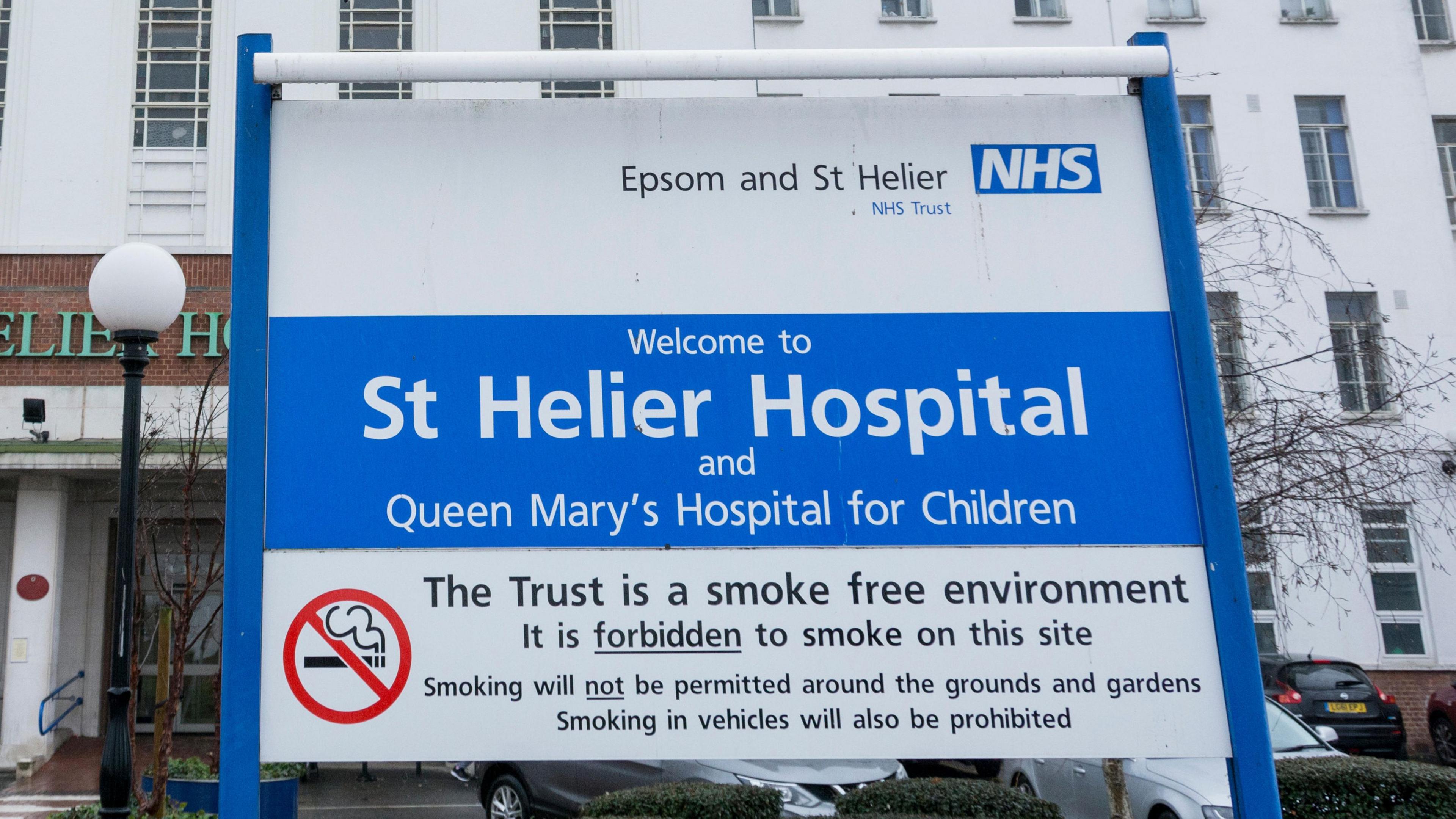 Exterior of St Helier Hospital shows white building entrance with blue and white Epsom and St Helier NHS trust sign welcoming patients and stating it is smoke free