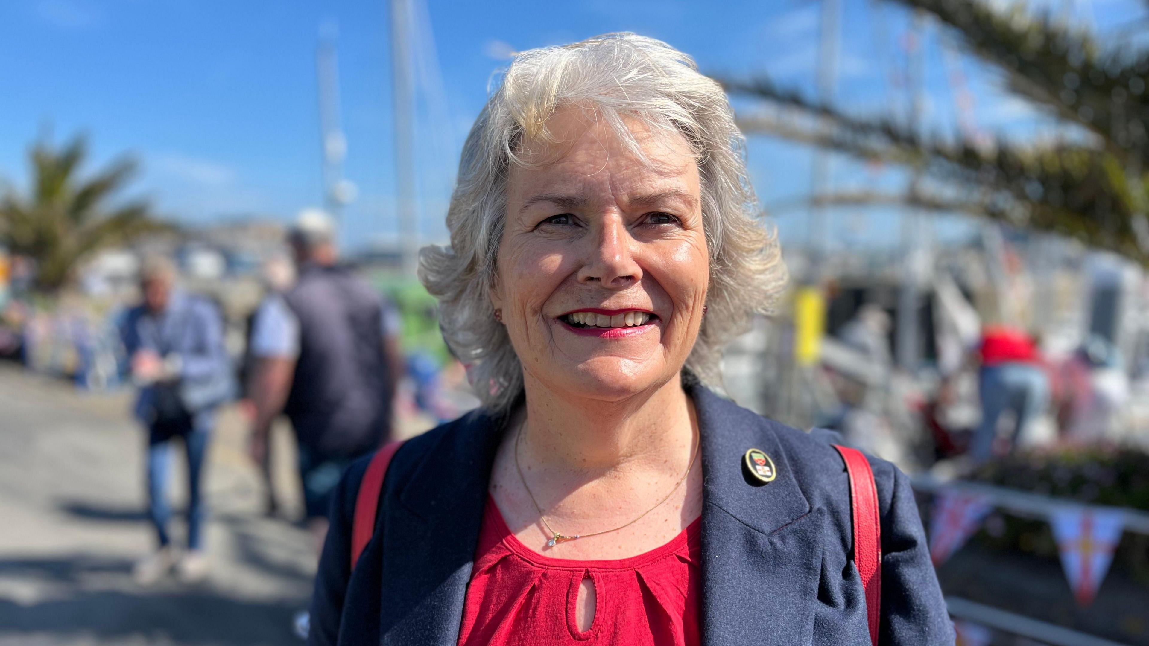 A picture of a smiling women with grey hair. She is wearing a red top, a navy blue blazer, and you can see the red straps of her backpack on her shoulders.