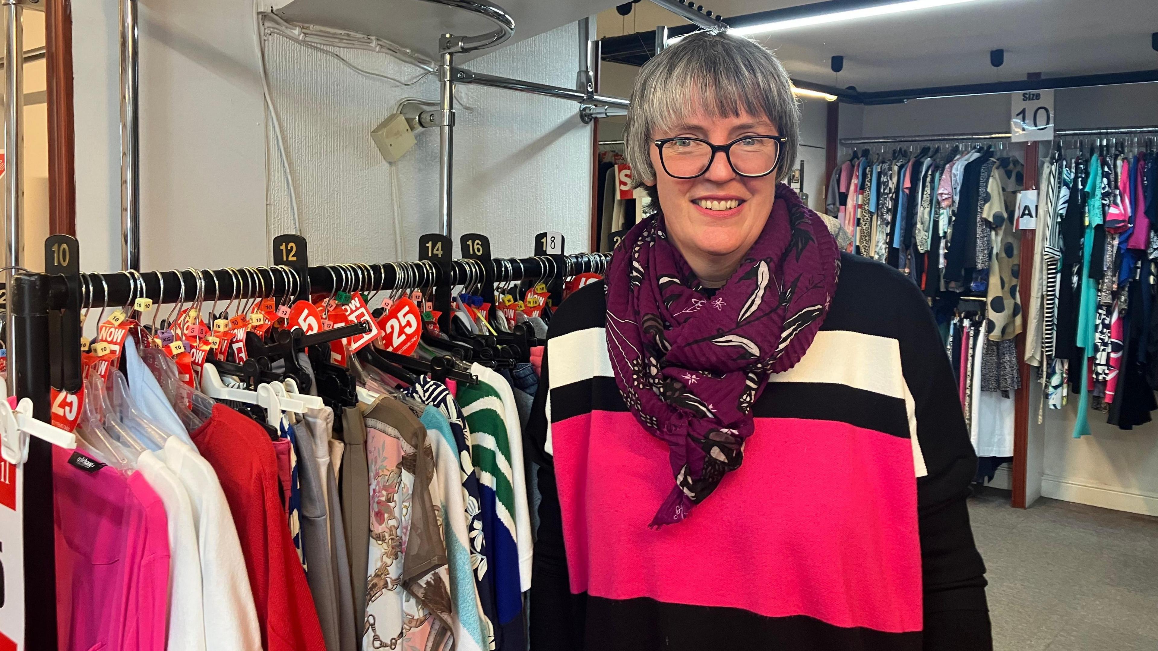 A woman in a pink, black and white top with a scarf around her neck and glasses is smiling at the camera. Behind her and to the left hand-side are rails of clothes
