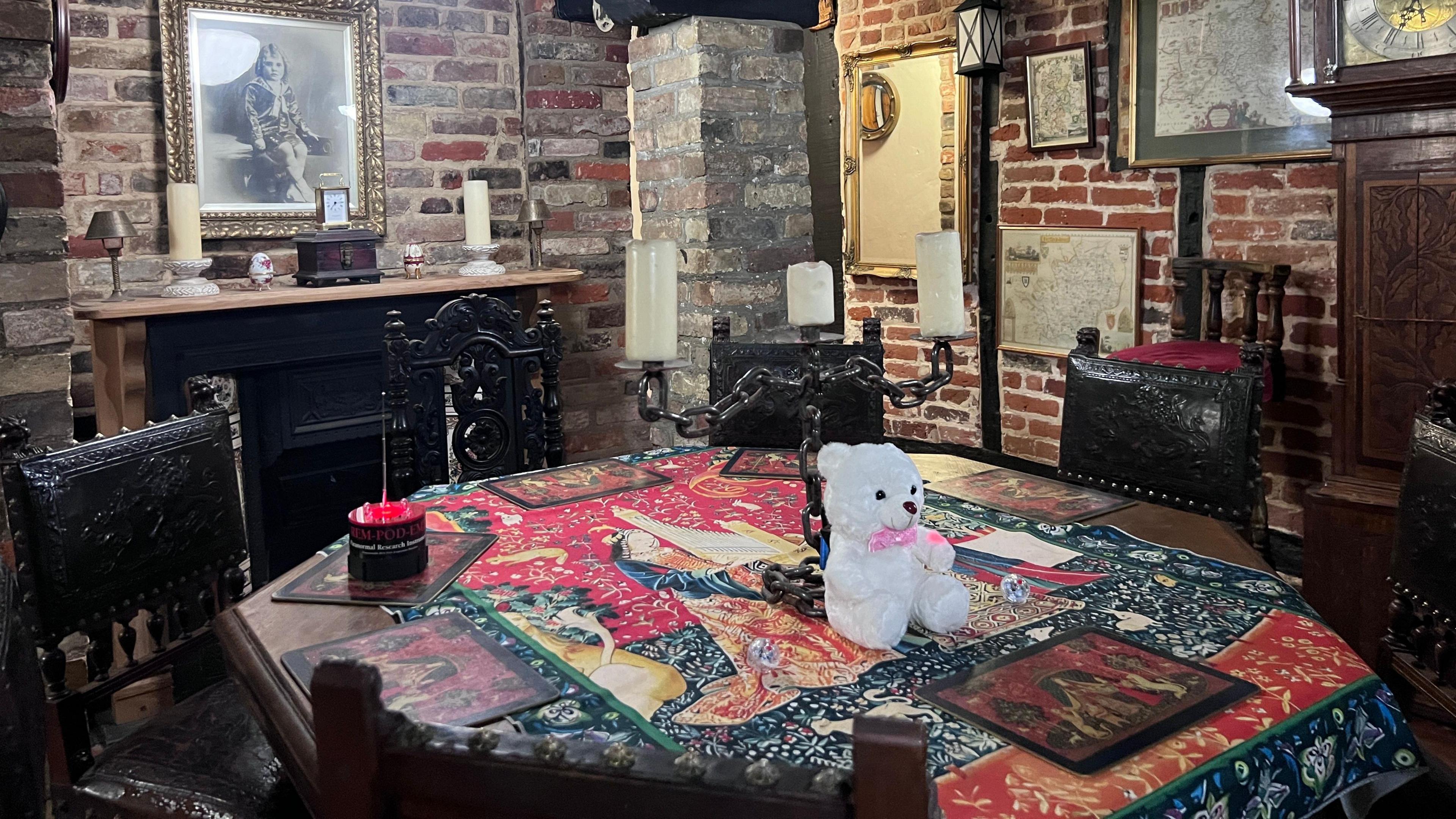 A dining table in a dark room. It has table mats on it, a bear ornament and is surrounded by dark wood chairs. The walls behind are brick and there is an open fireplace with candles on the mantle