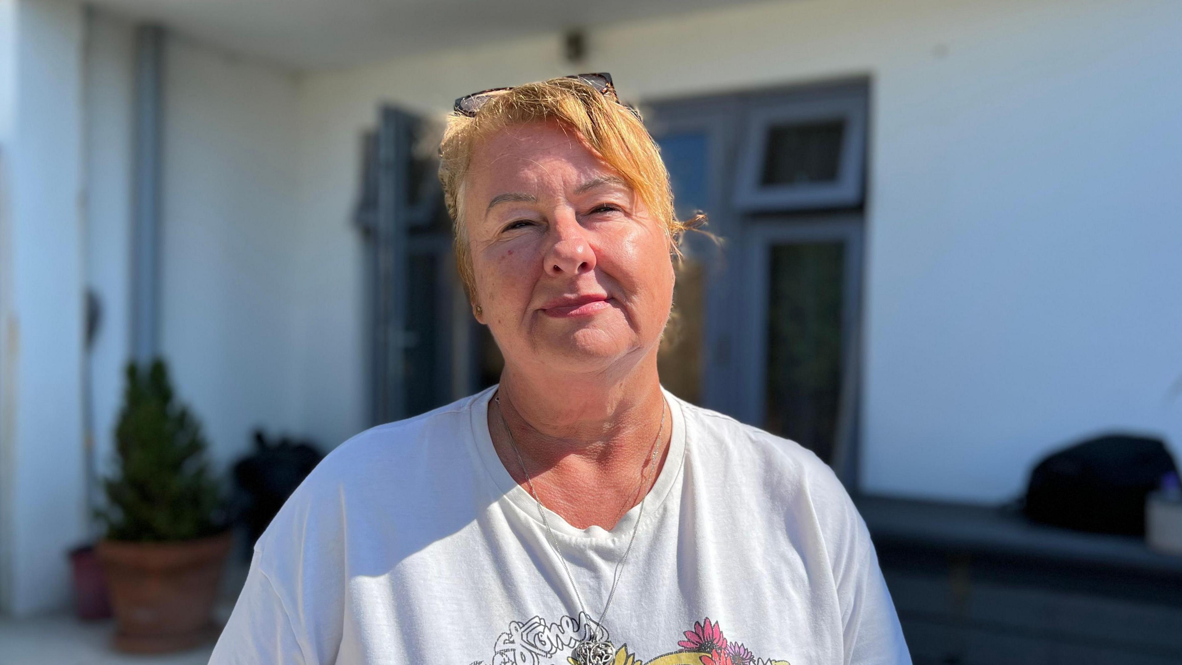 Annette is a woman with light strawberry blonde hair. She is wearing a white t-shirt and has glasses on her head. She has a silver necklace around her neck. She is standing in her garden, in front of the backdoor to her living room. There is sunshine on her face and she is looking into the camera.