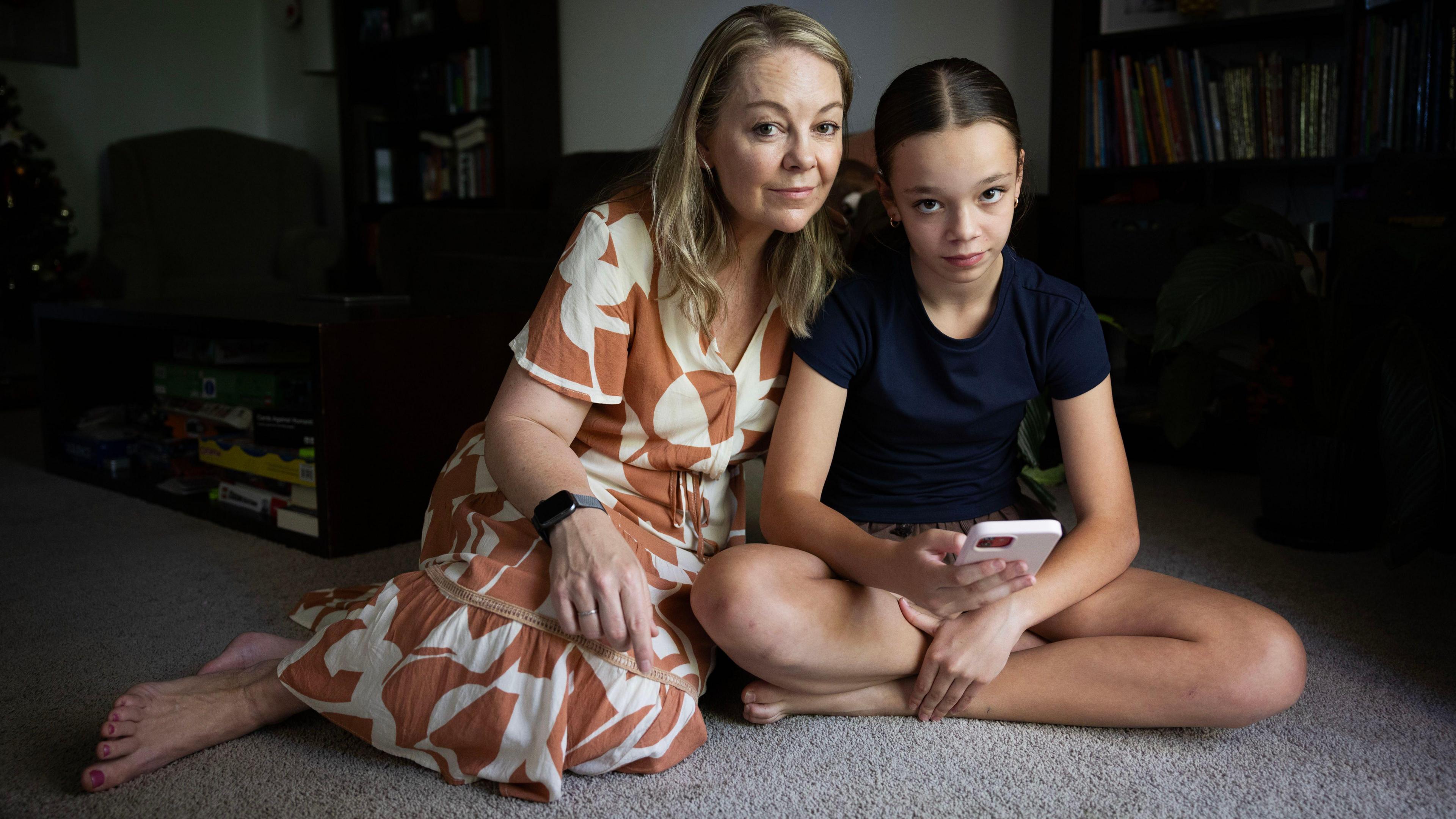 A  woman in a brown and white dress sits on a carpeted floor with her teenage daughter, who is holding a pink phone