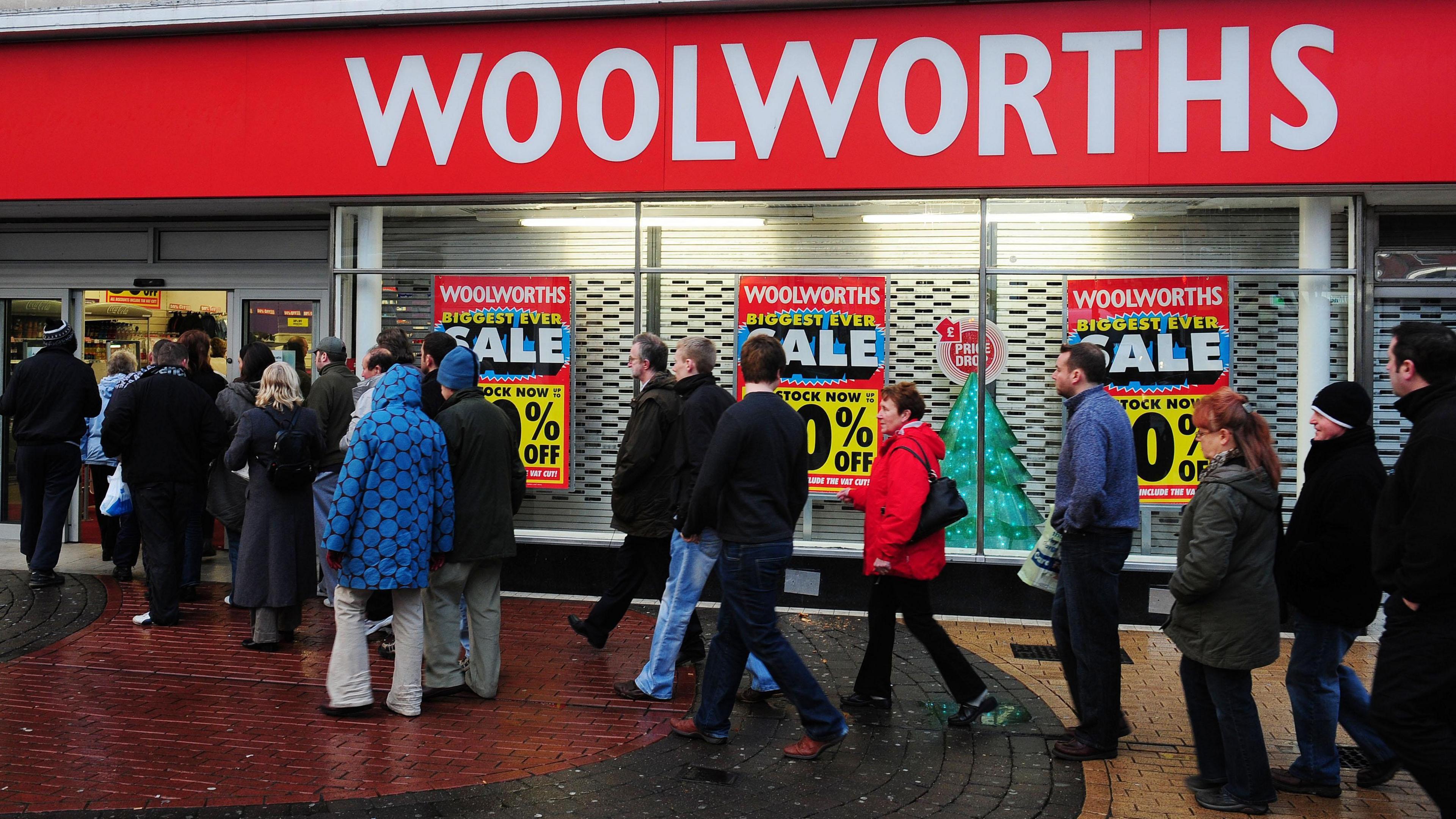 People queuing to enter a Woolworths store, which has a large red banner across its front that says "Woolworths" in white writing. The shopfront has large posters advertising a "biggest ever sale" at the shop. The people queuing are mostly wearing coats on a cold day.