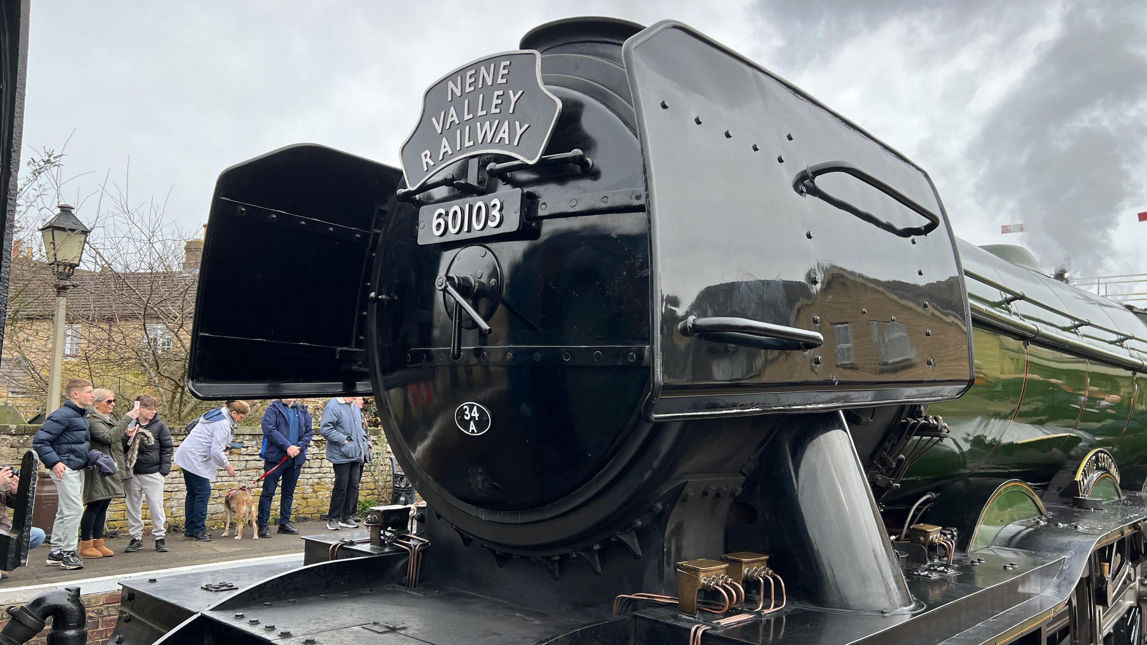 The front of the Flying Scotsman. A crowd of people on the other side of the platform are looking and taking photos.