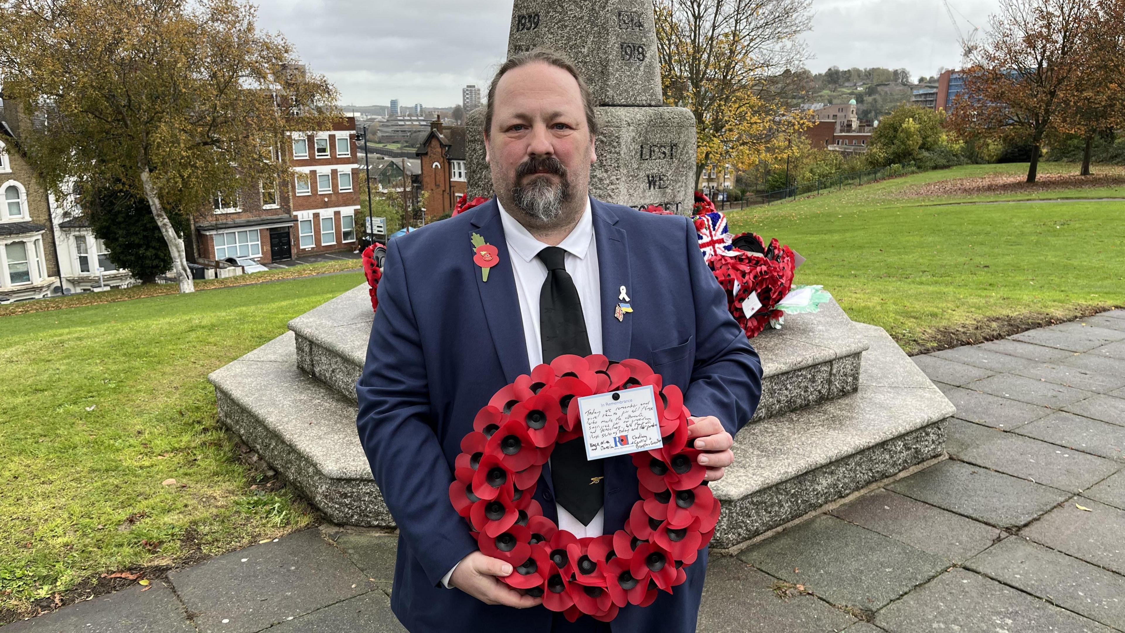 A man with goatee beard wearing a blue suit laying a wreath of popies at a war memorial on a hill.