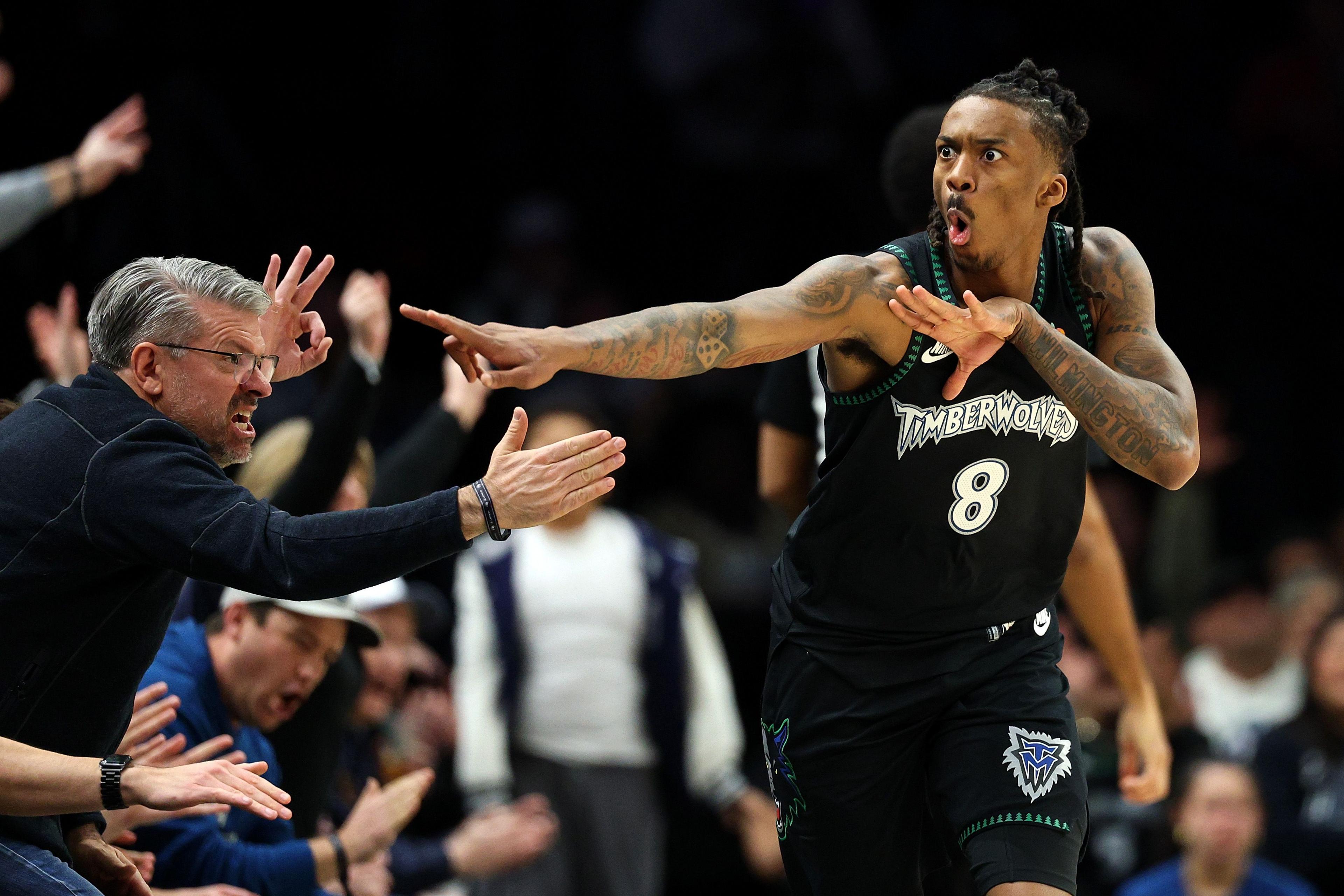 Basketball player Bones Hyland, in a black Minnesota Timberwolves uniform, celebrates near the sideline, extending one arm outward and the other across their chest, while fans reach out enthusiastically from the front row.