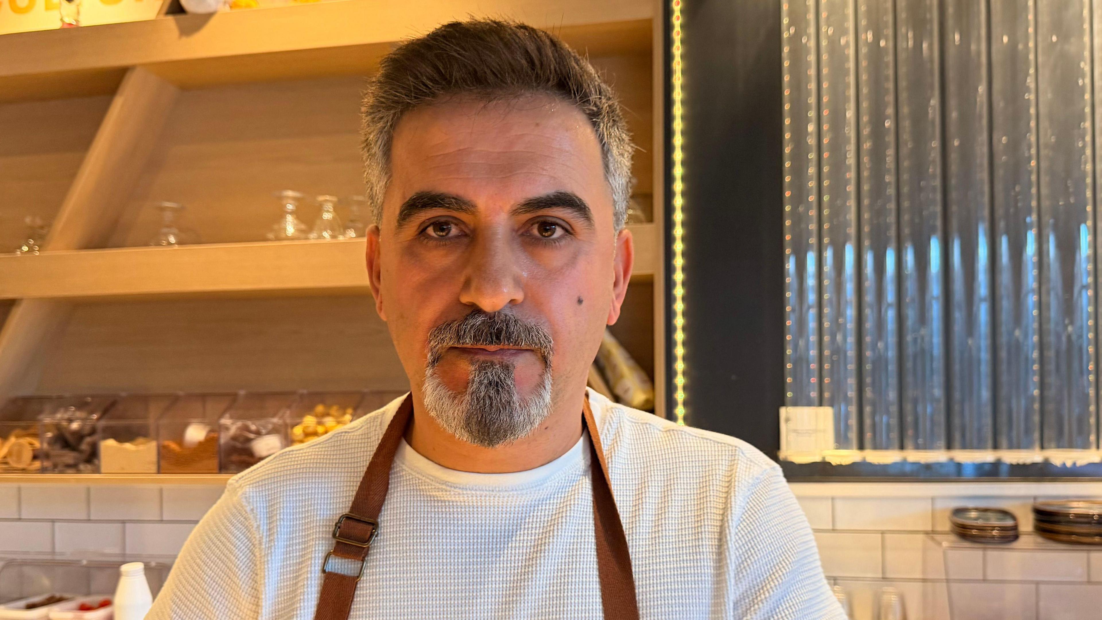 Yilmaz Aydemir wearing a white long-sleeved shirt and an apron. He has a grey and white shaved beard and moustache and dark thick eyebrows. His eyes are dark and he is standing behind the counter at a modern dessert parlour. 