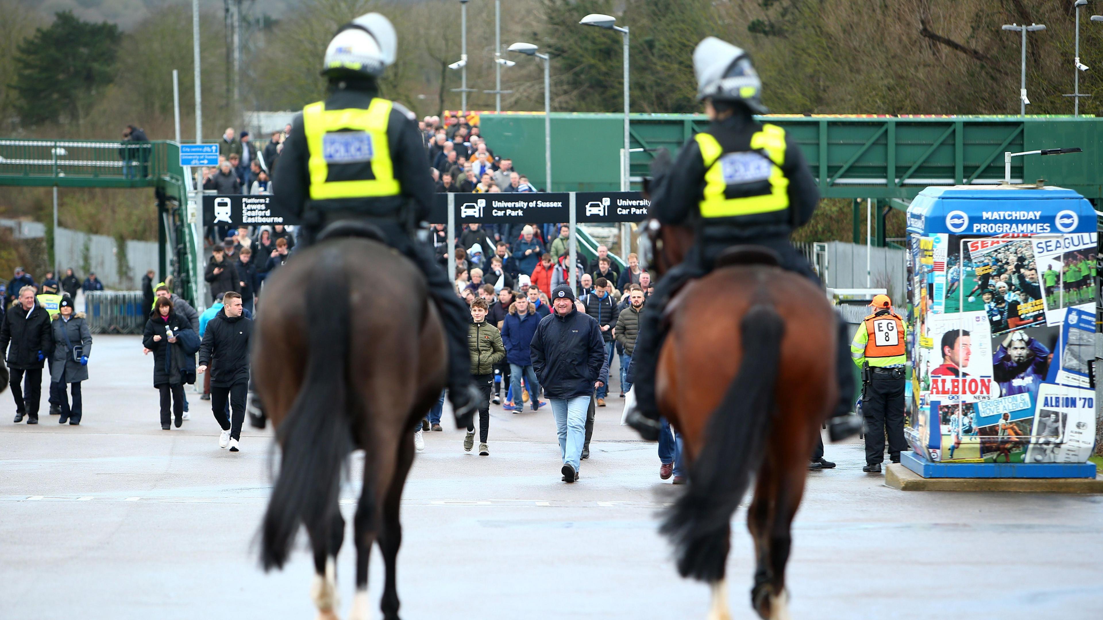 Two police officers wearing helmets and high-visibility jackets sat on top of horses. There is a large crowd of football supporters walking towards them and a matchday programmes vendor to the right. 