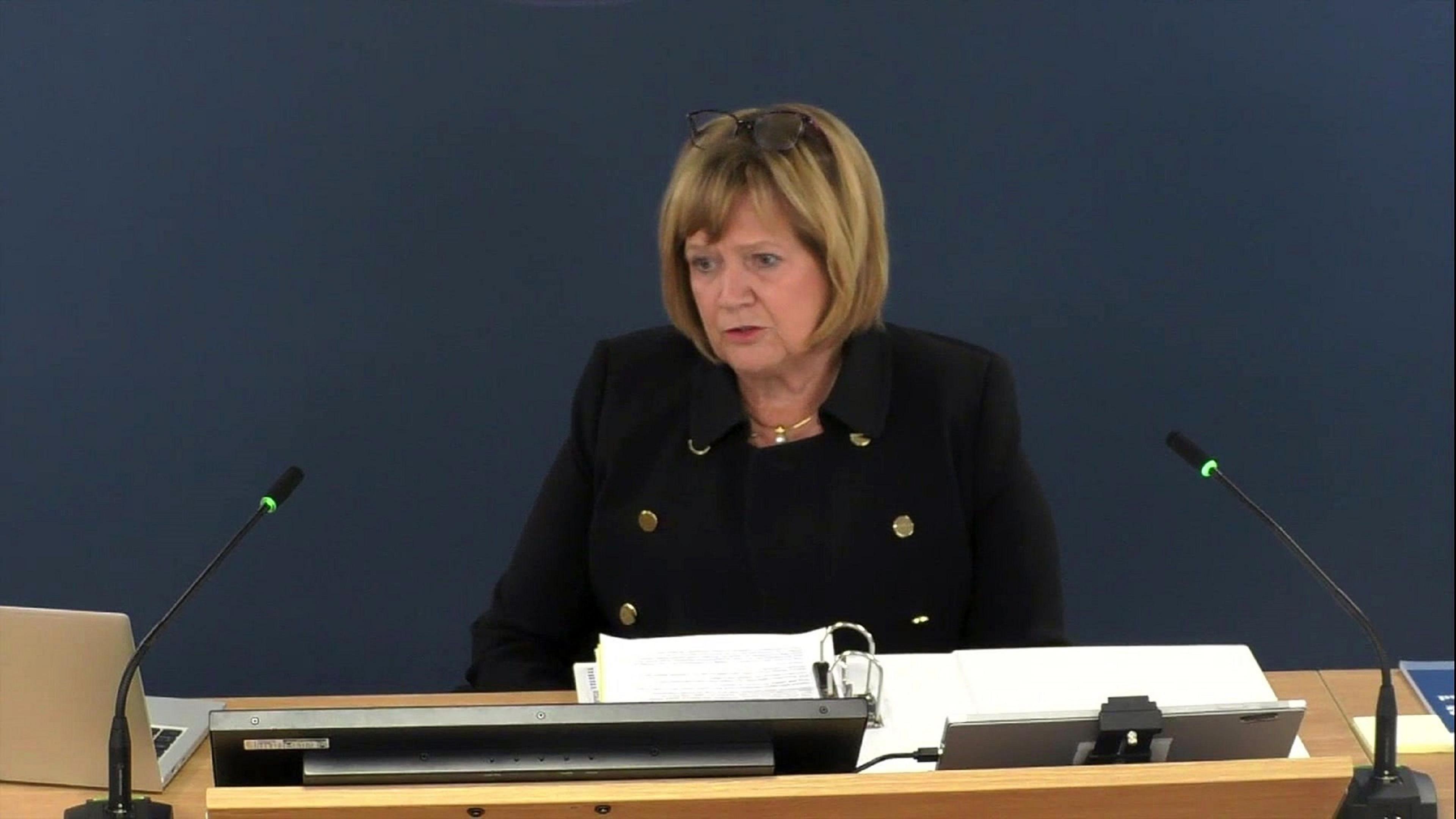 Baroness Hallett has blonde bobbed hair. She is wearing a black jacket and sitting behind a bench with computer screens and two microphones