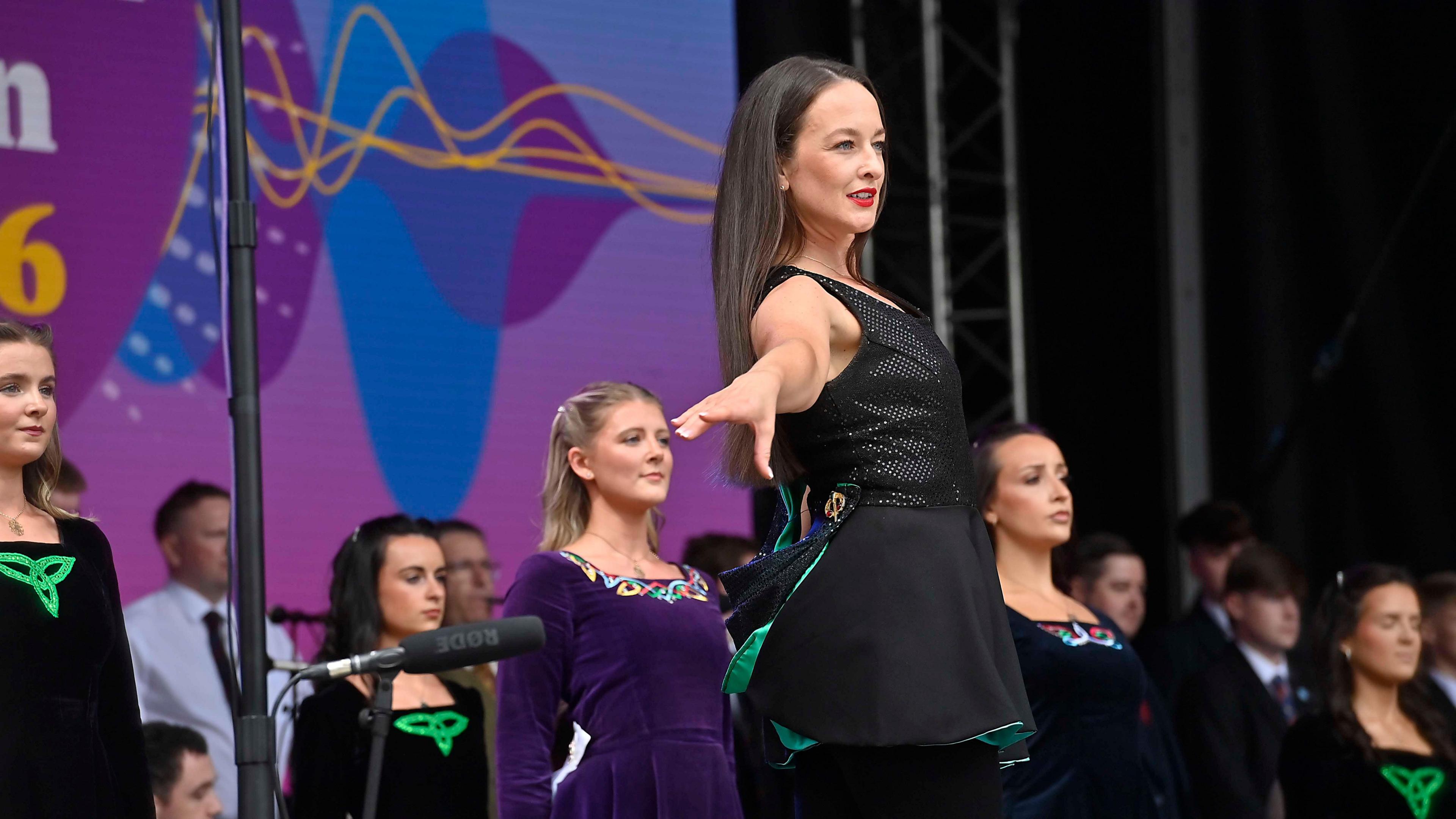 A woman wearing black clothing stands with an arm outstretched. She has long dark hair, and there are more people behind her, also wearing dark clothing. 