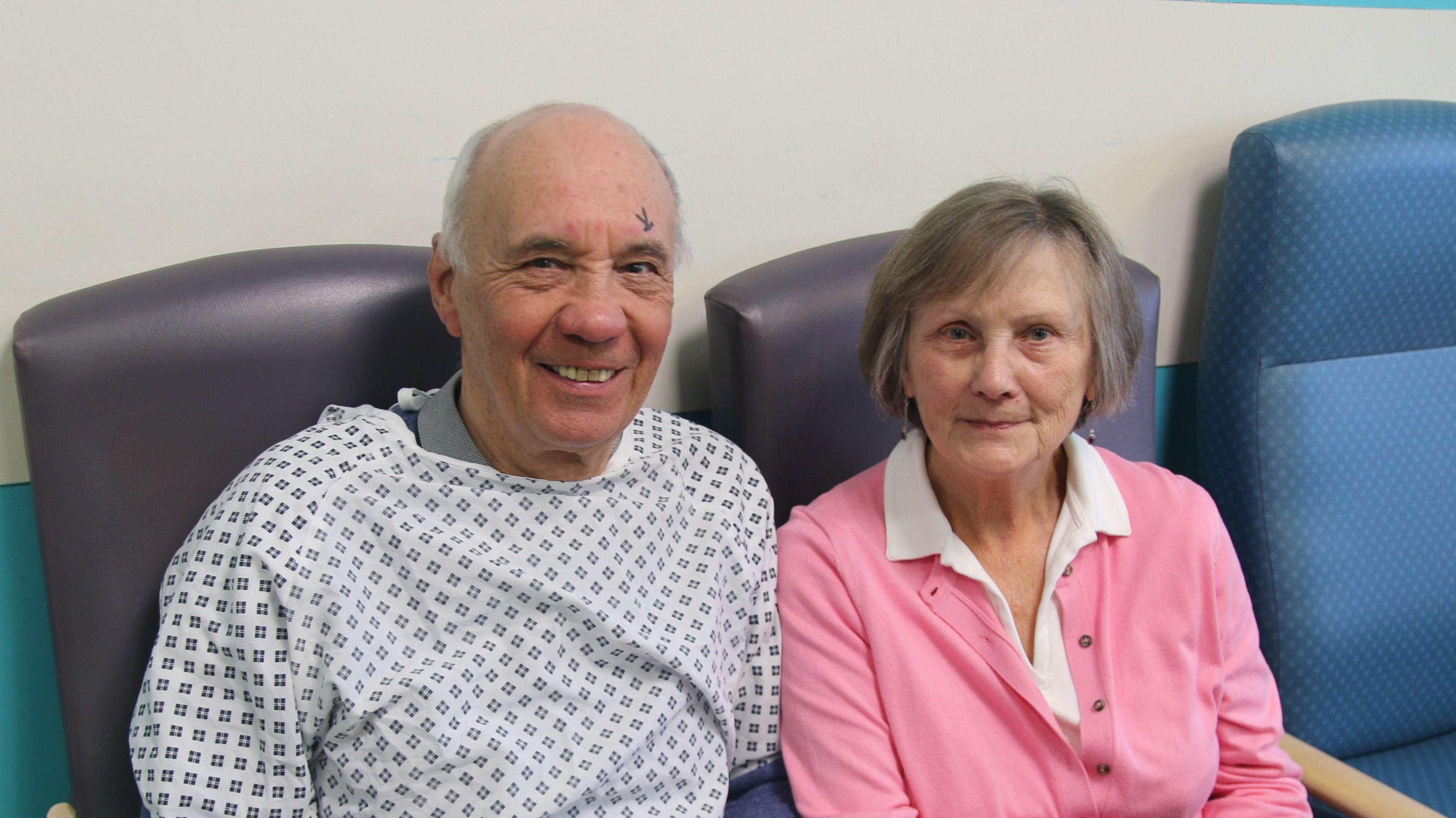 John and Freda McBean sitting next to each other in purple hospital chairs. Mr McBean wears a hospital gown and appears to have a surgical pen mark over his left eyebrow, while Mrs McBean wears a pink cardigan and white shirt