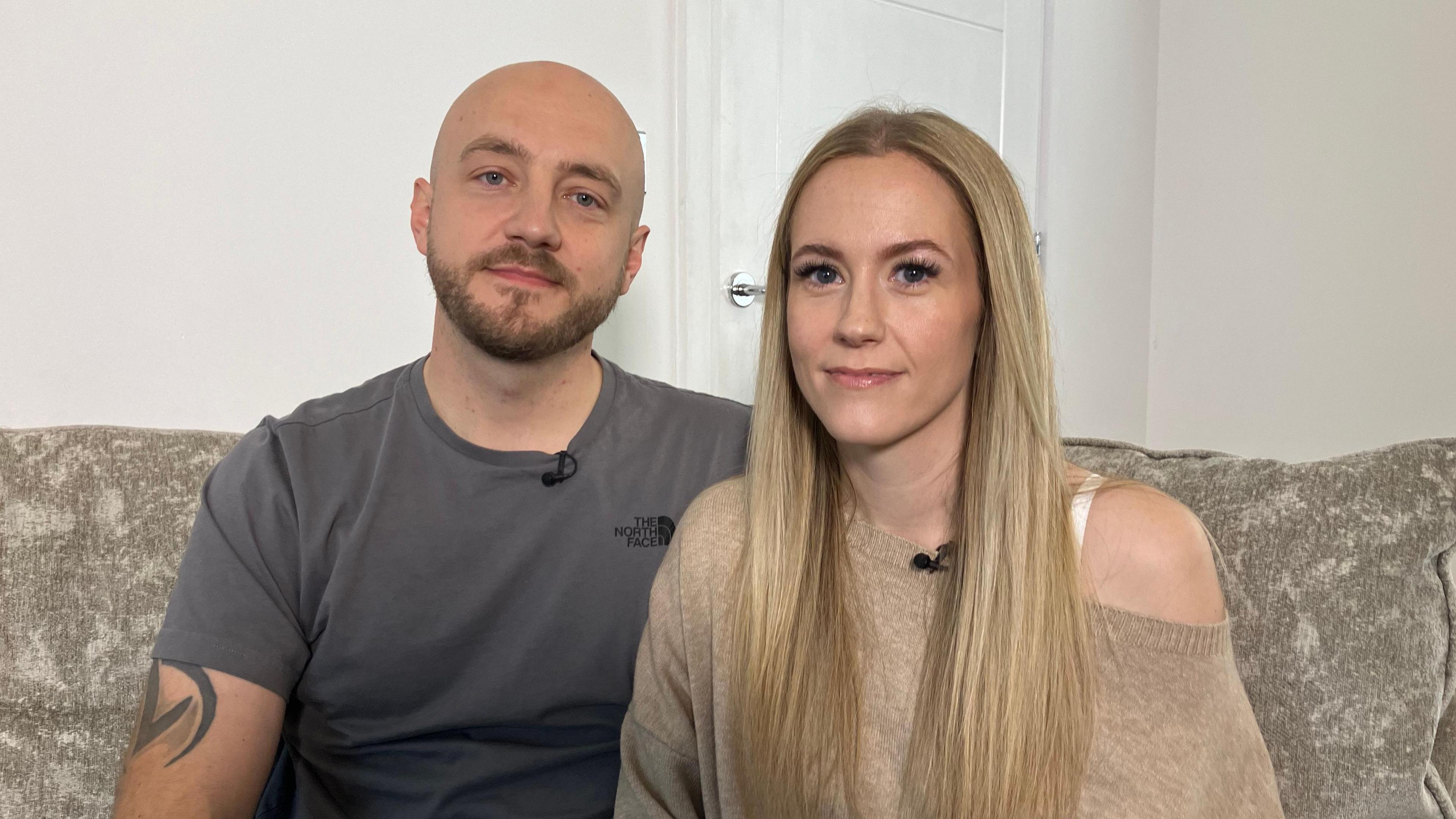 Tom and Becky Williams are sitting on a large, neutral coloured sofa. Tom has a tattoo on his arm and is wearing a dark grey top, he is bold and has a beard. Becky is wearing a neutral coloured top, she has blonde hair and together they are looking at the camera. Being them is the door to their living room.