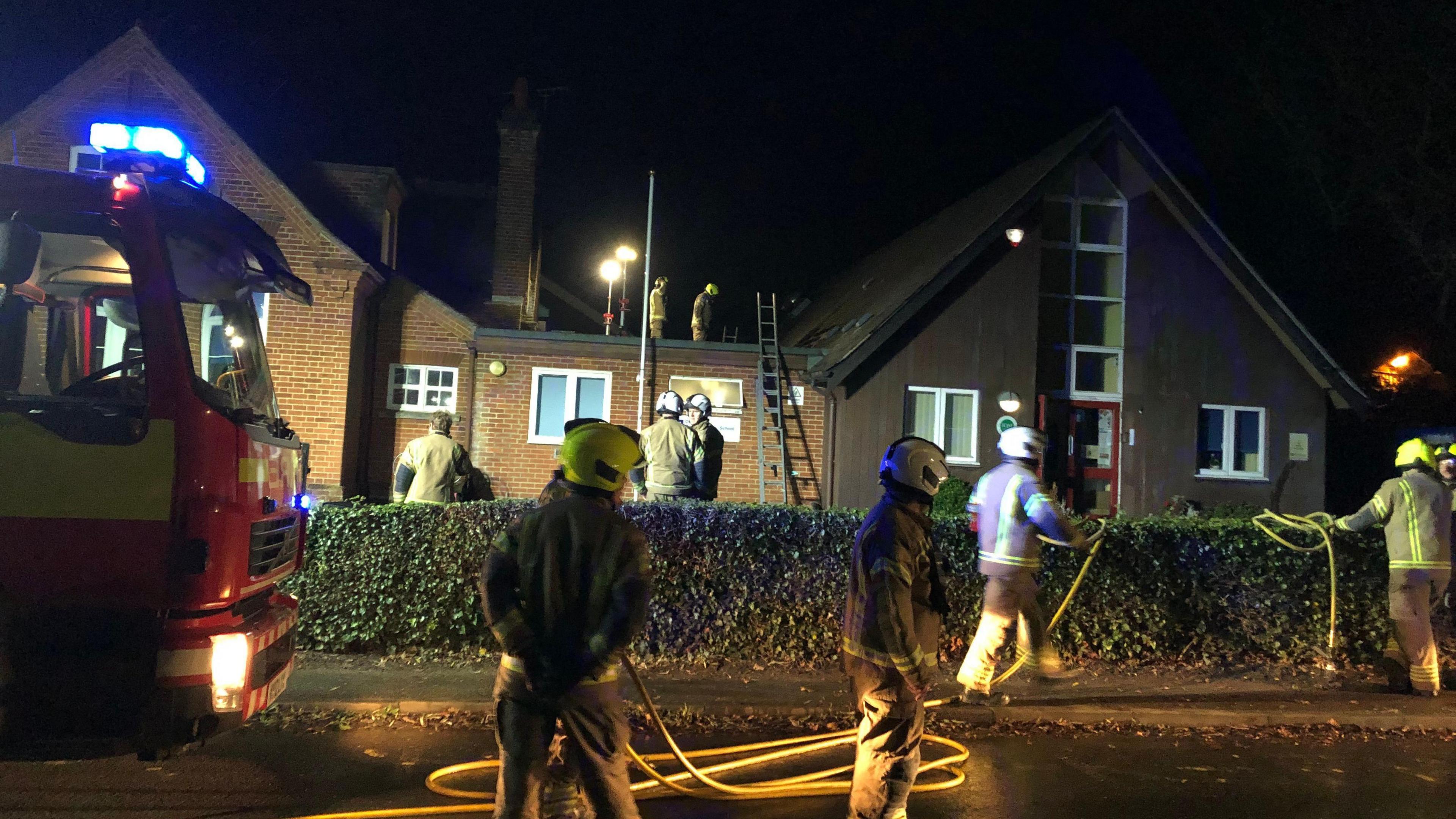 Firefighters surround the school with hoses and ladders. The front of a fire engine can be seen to the left of the photo.