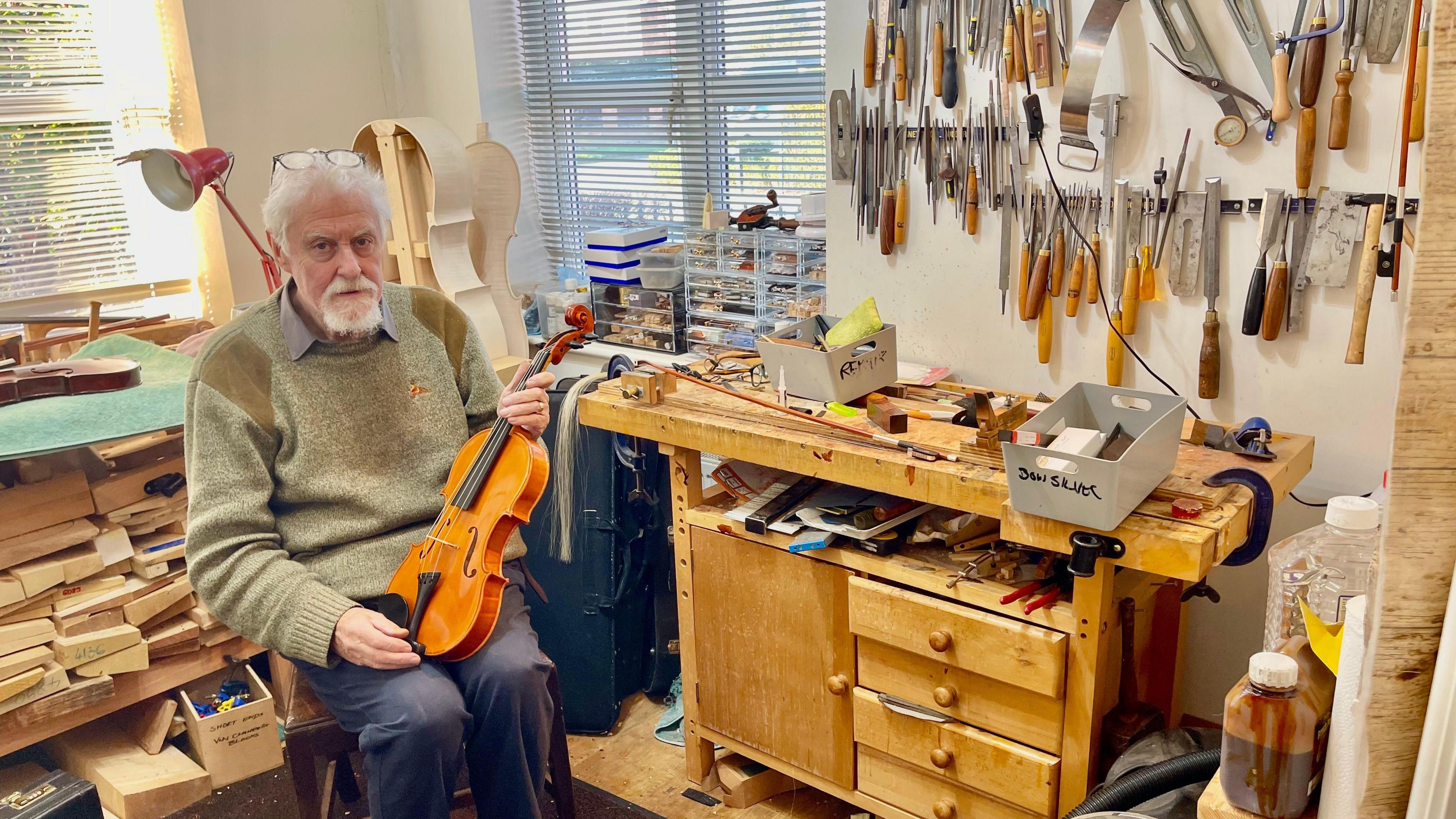 A man sits in a chair holding a violin in a well lit room of a house. The room is filled with a workbench, instrument cases, tools, wood, and instrument parts. Many tools are also hung from the wall. The man has short grey hair, a grey beard, glasses balanced on his head, a green jumper with brown shoulder pads and navy trousers.