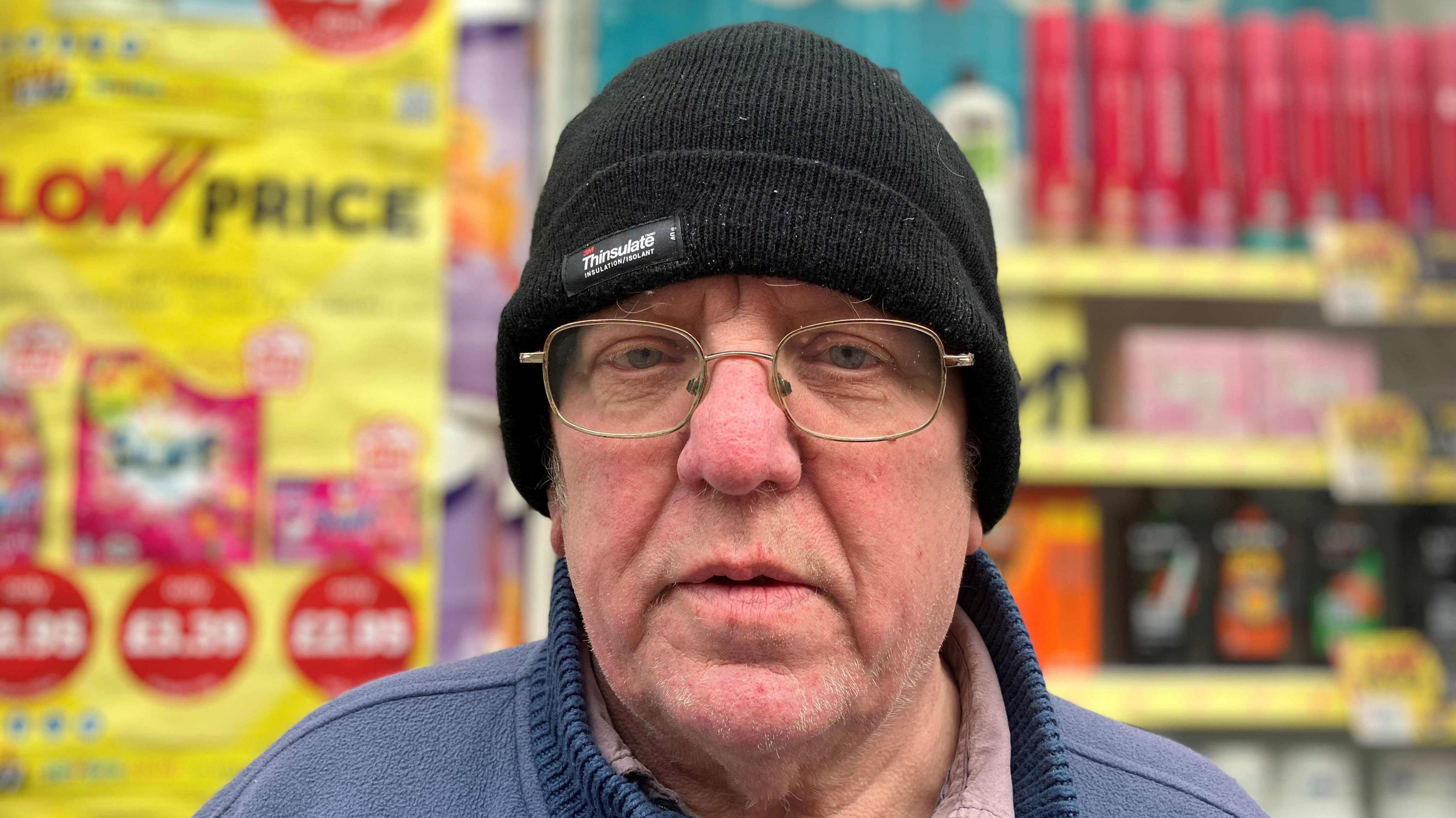 Graham Charles looking at the camera, has a black beanie hat on and glasses. He is wearing a blue jumper.