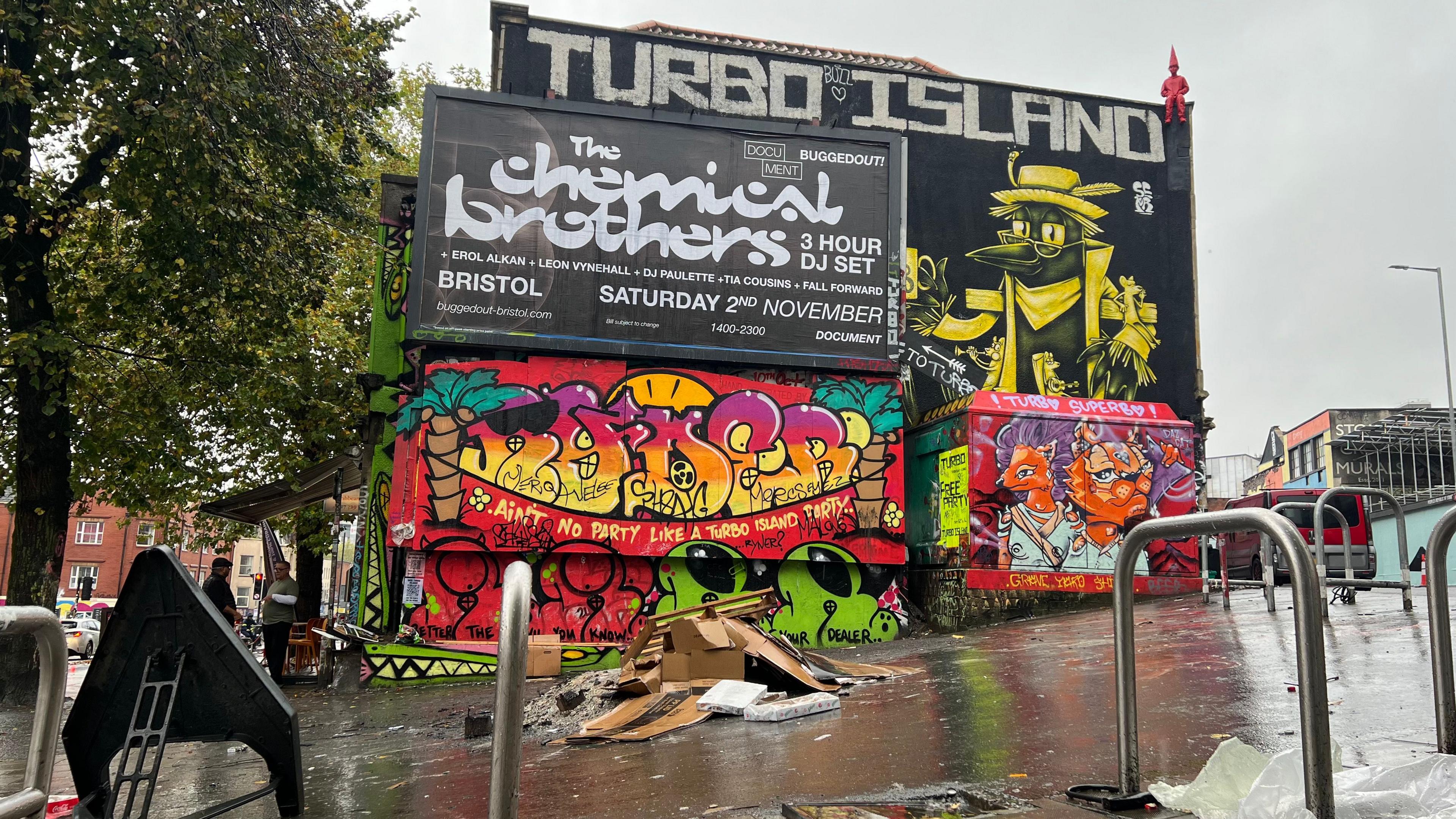 A wide shot of Turbo Island in Stokes Croft in Bristol showing the remains of a fire in the foreground and large walls with graffiti and posters on them in the background