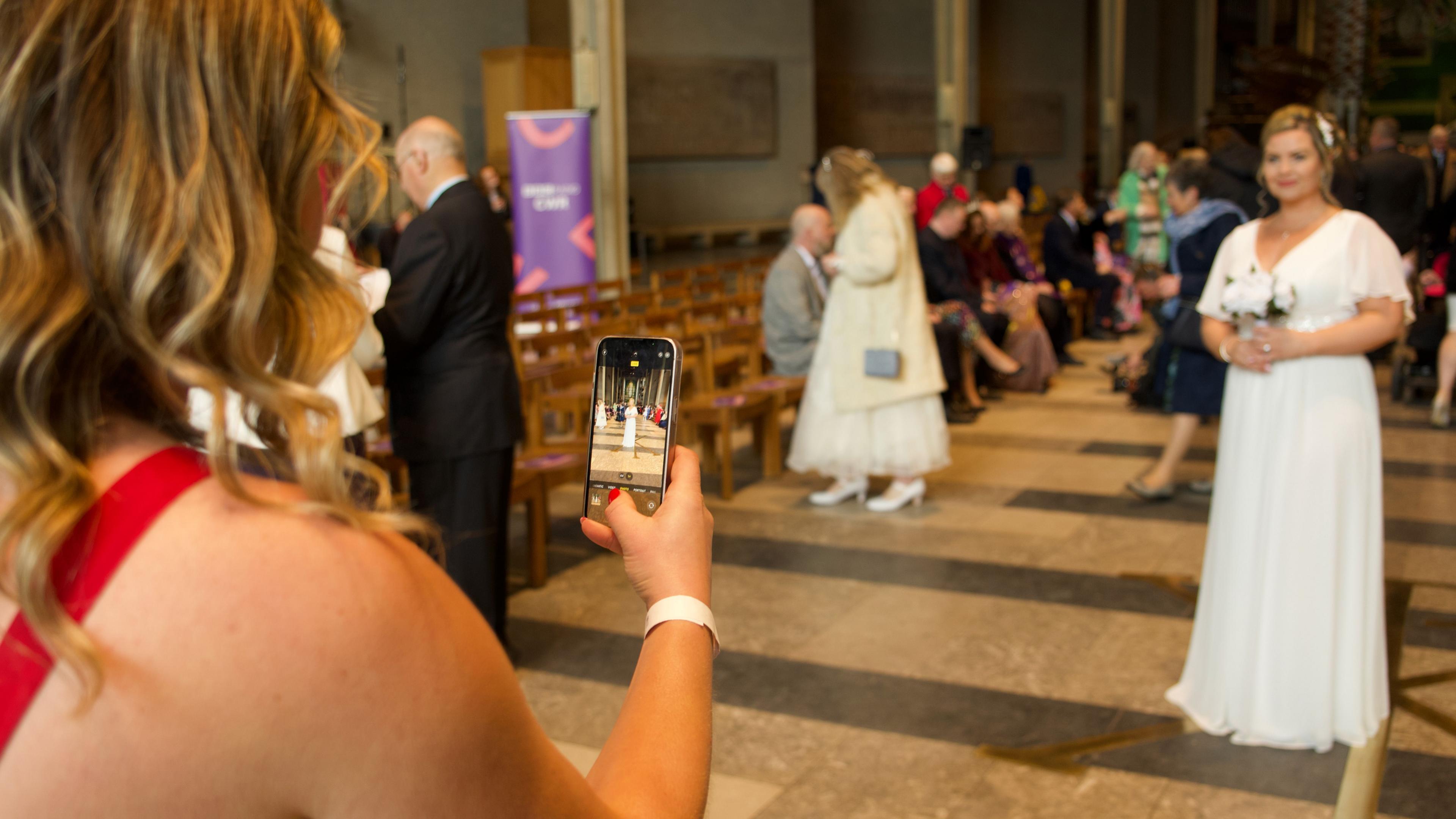 A woman takes a photograph on her smartphone of a woman in a white wedding dress