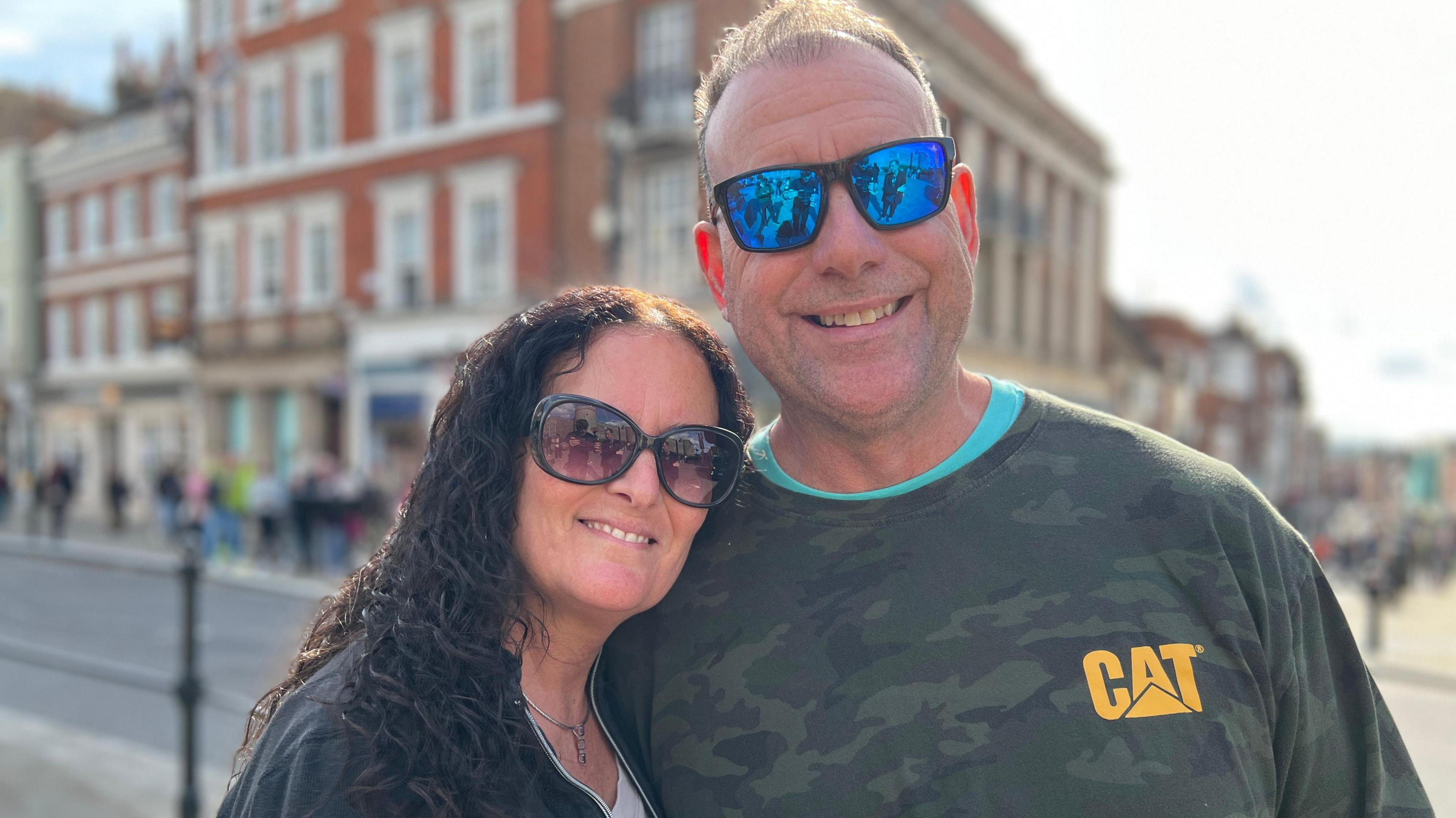 Annette Wagenhofer and her husband Gerard right both are wearing sunglasses and smiling at the camera with a street behind them with red brick buildings 