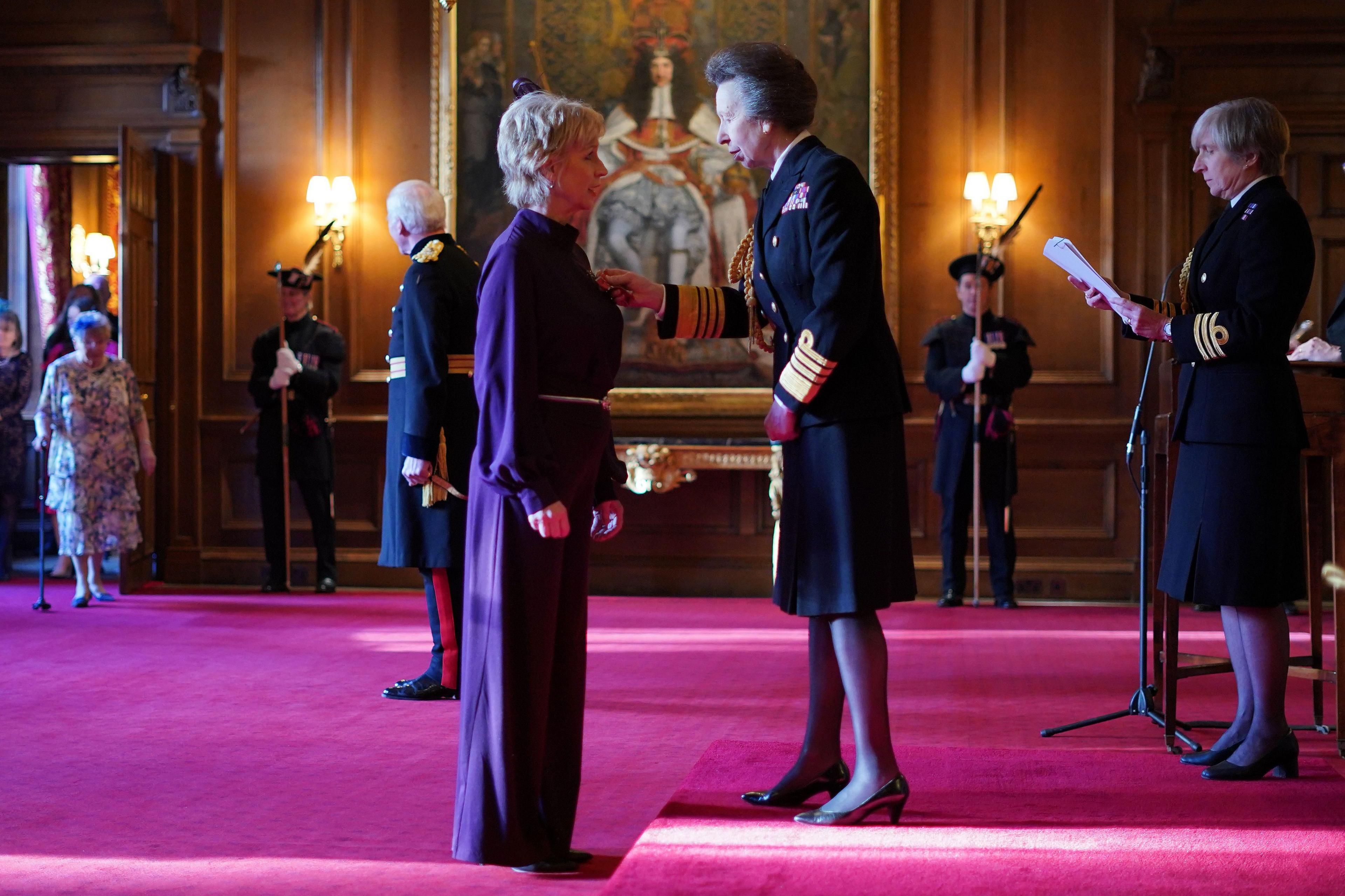 Sally Magnusson in a long purple dress, is standing on the left in profile speaking to the Princess Royal. The Princess is standing on a slightly raised platform and is in a Royal Navy uniform. The carpet is light purple, the walls wood-panelled and there are three uniformed guards in the background.