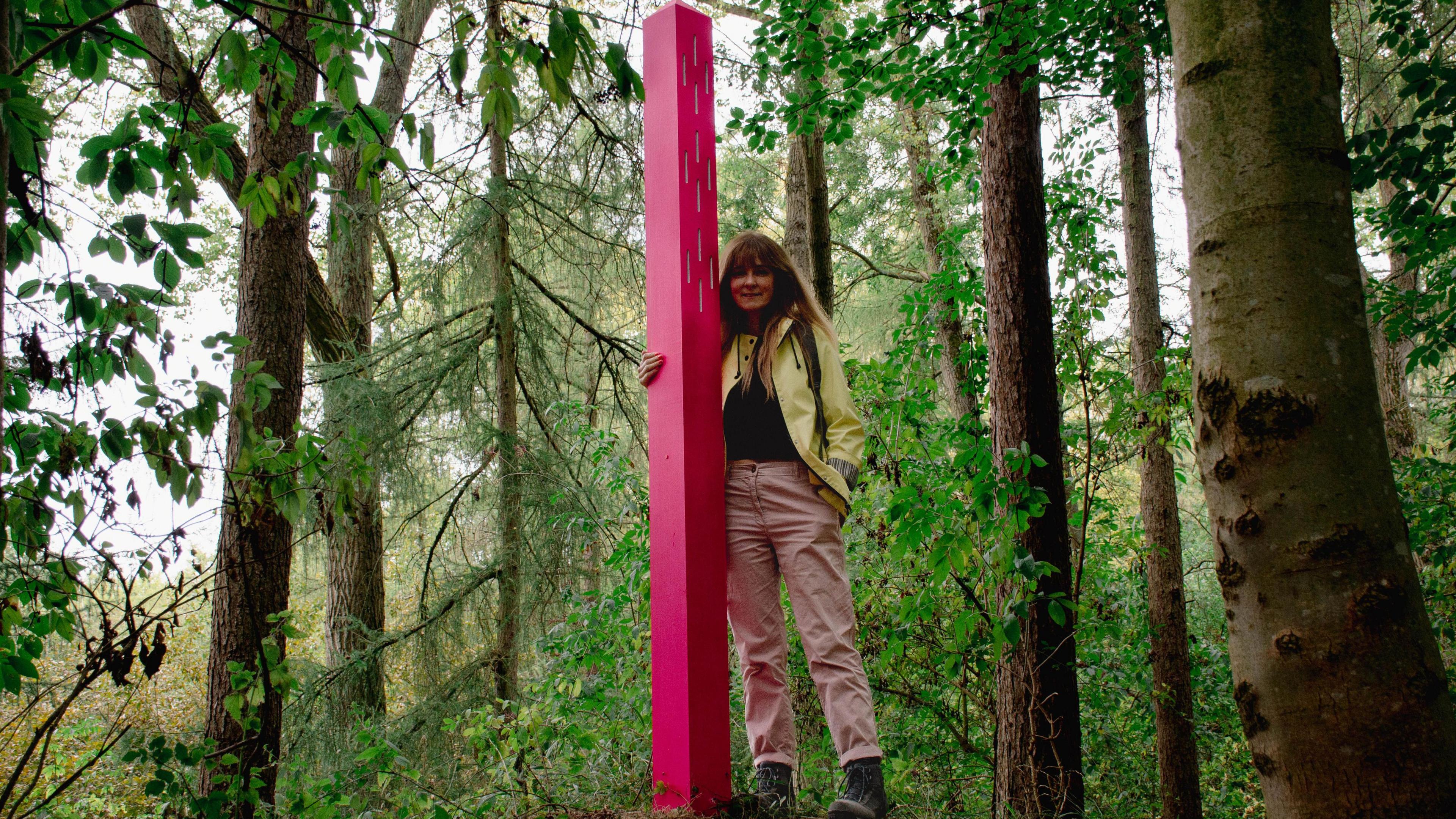 Artist Beccy McCray, with long brown hair, wearing a light-coloured jacket and trousers, stands next to a pink waymarker amid tall trees.