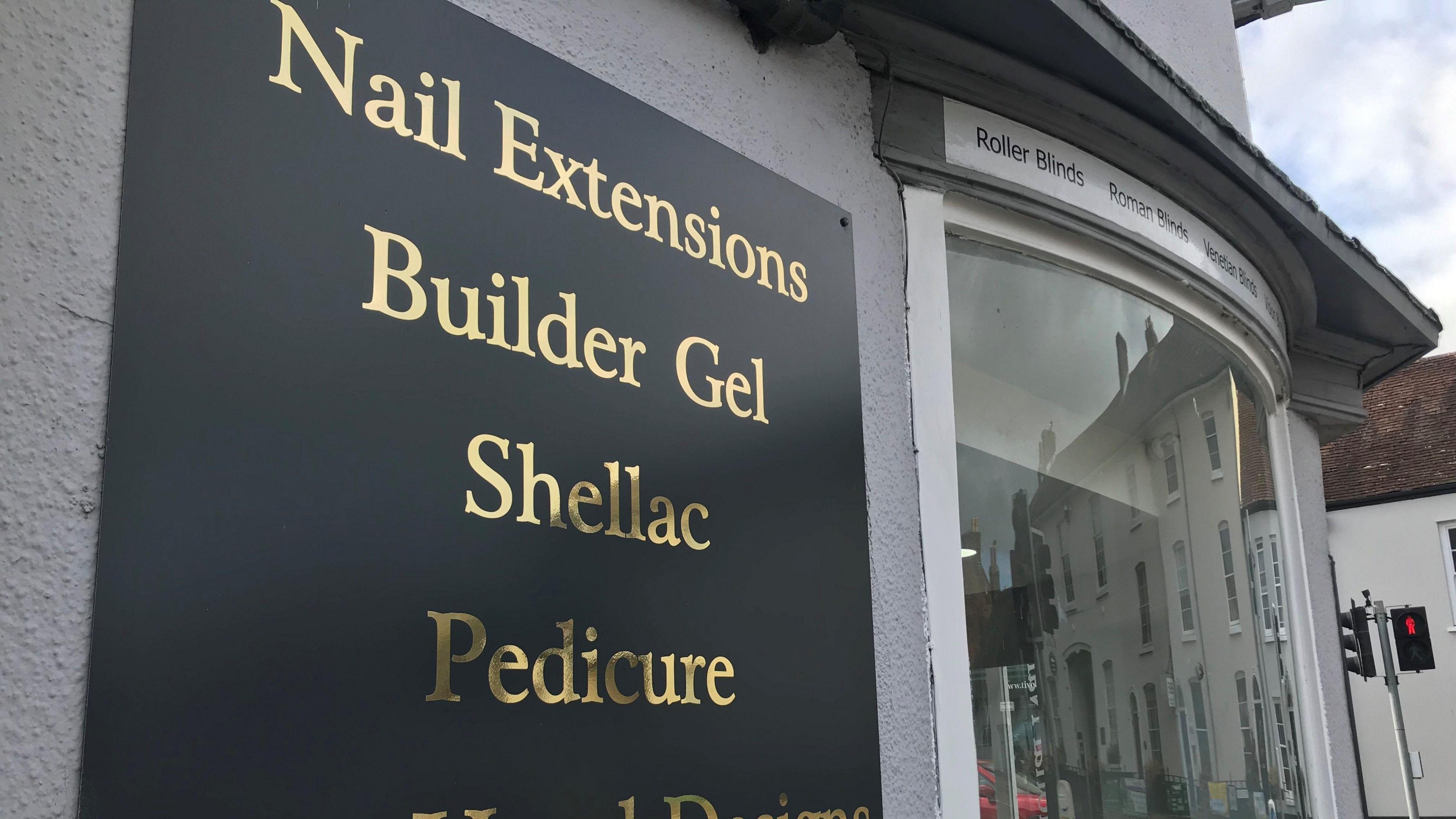 Another view of The Nail Room shows a square black sign which lists salon services like nail extensions, builder gel, shellac, pedicure all in gold lettering and a bay window to the right of the frame