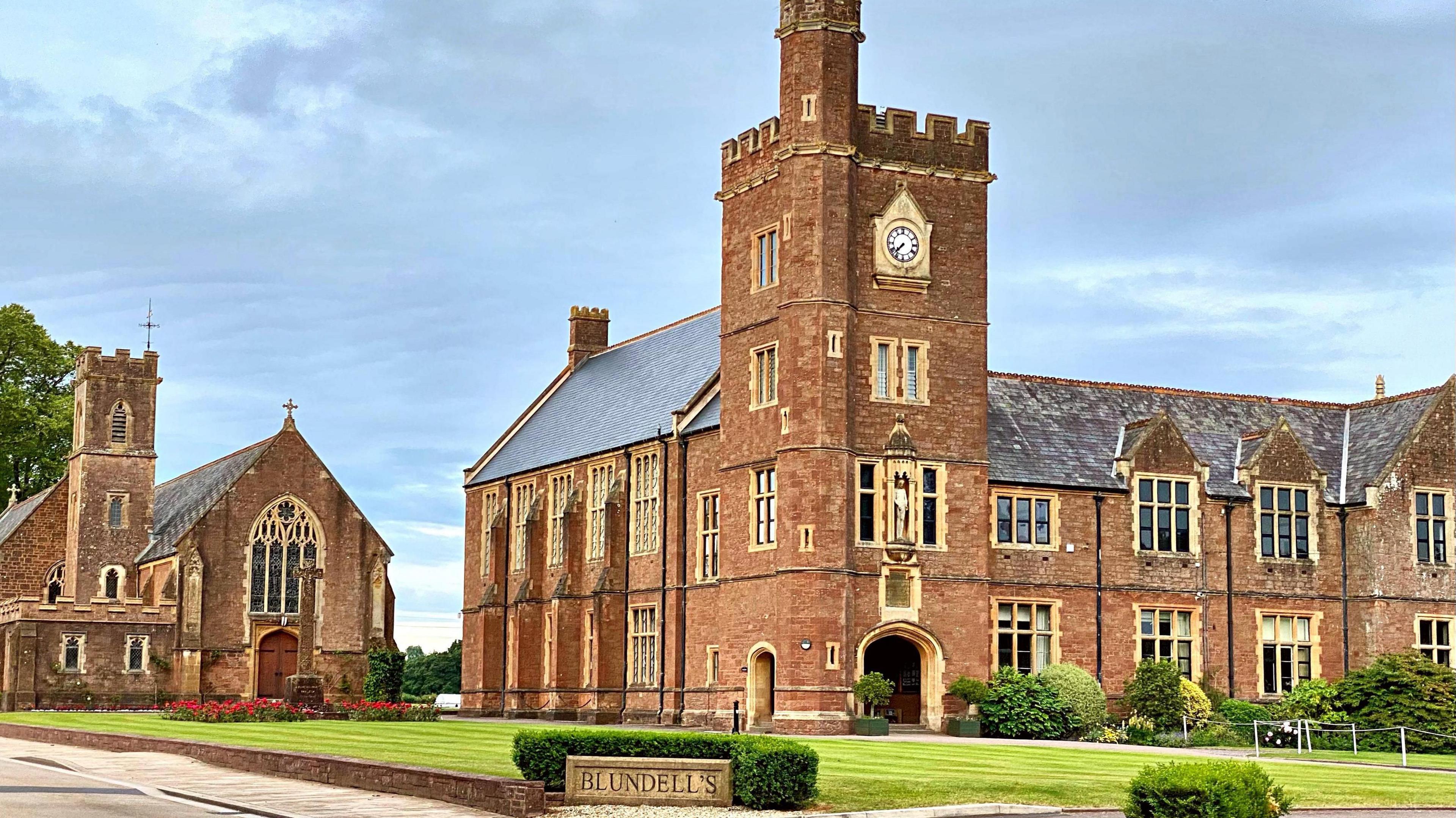 Blundell's School in Tiverton, with several brick buildings with a lawn and bushes outside it and a stone sign with the school's name on it in front of a bush.