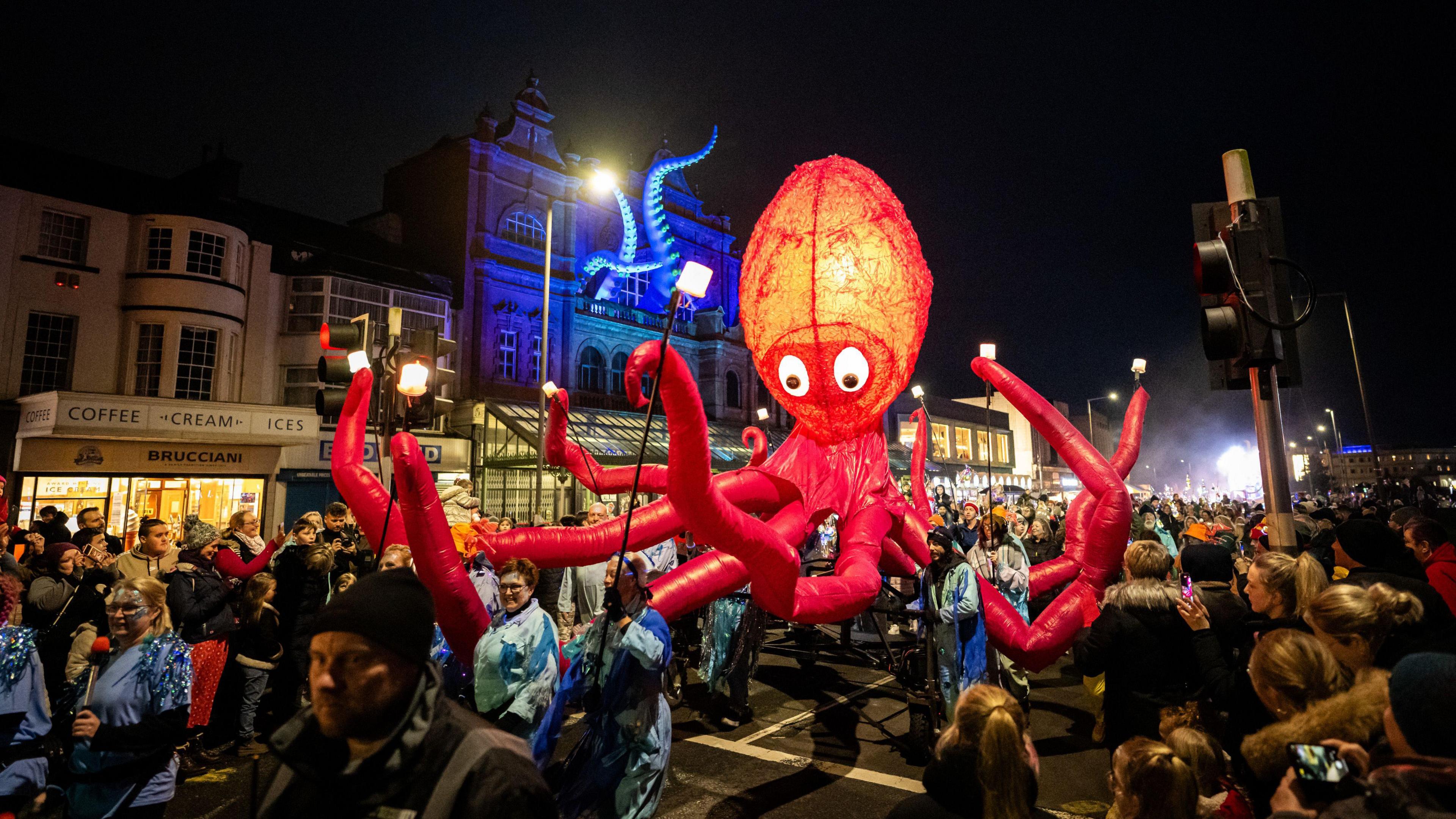 A red giant inflatable octopus with a glowing head being paraded through the streets of Morecambe surrounded by people.