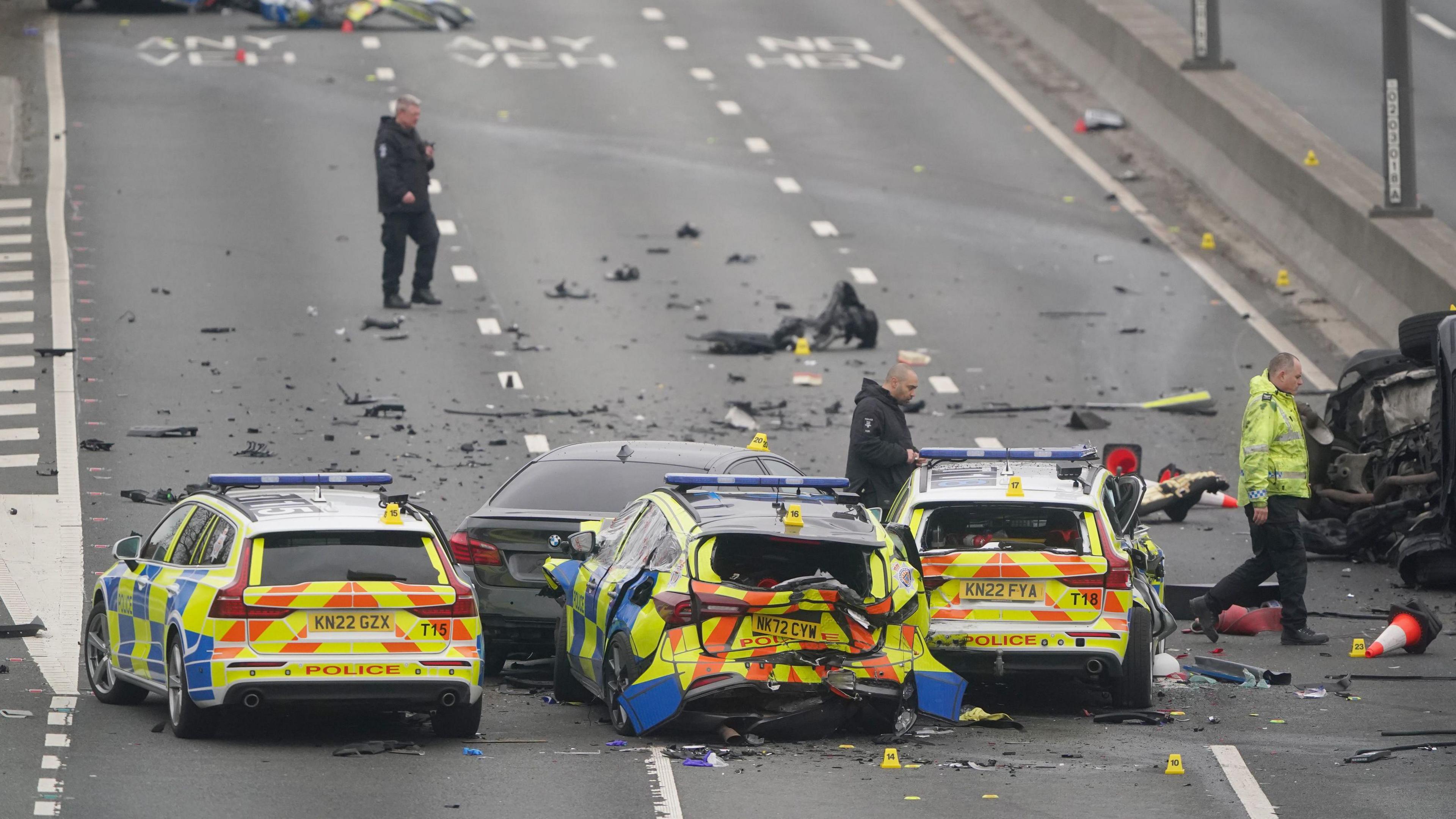 Three police vehicles, smashed with debris all over three lanes of an A road. A black BMW can be barely seen in front of the middle police car. Three men walk across the road, assessing the scene, in police uniforms.
