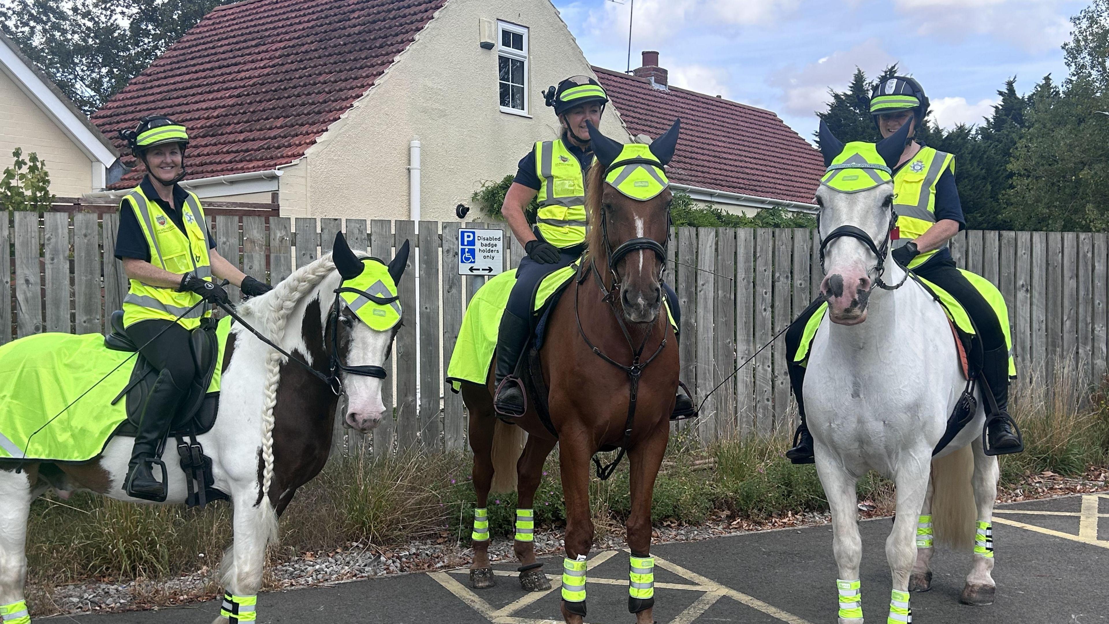 Three horses in high vis, the horse on the left is white with dark spots and a braided mane, the central one is large and brown and the horse on the right is white. The women riding are also wearing high vis and head cams.