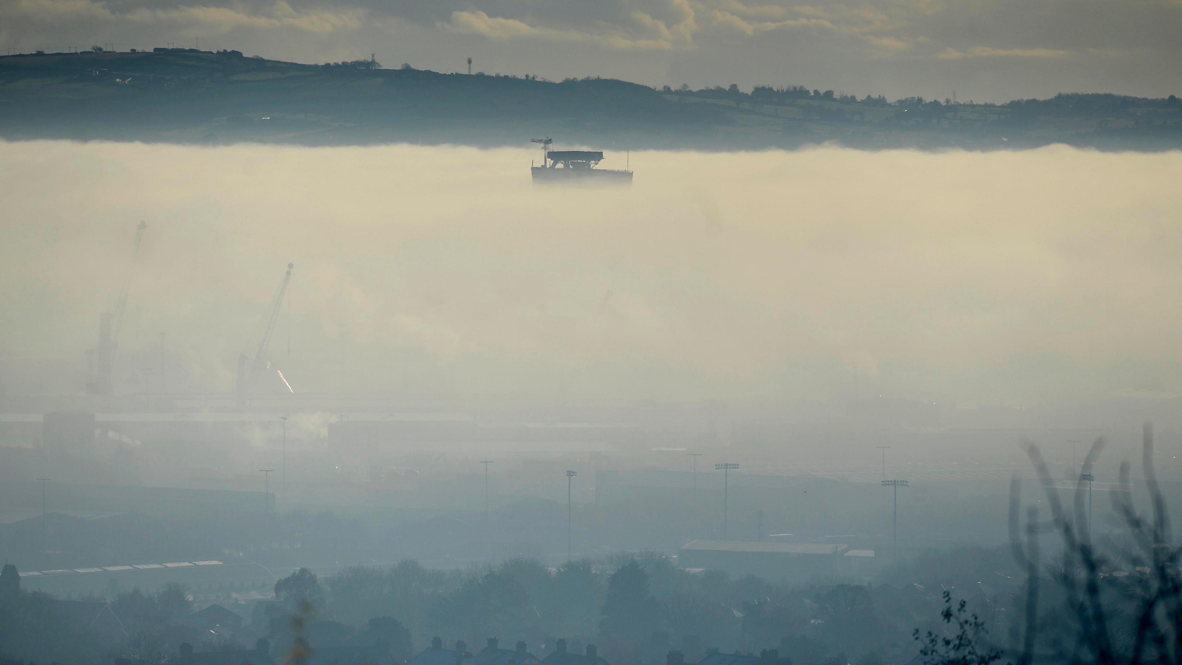 A still, wide image showing a thick layer of fog covering most of Belfast. Sitting above the cloud of fog is the top of the yellow Harland and Wolff cranes.