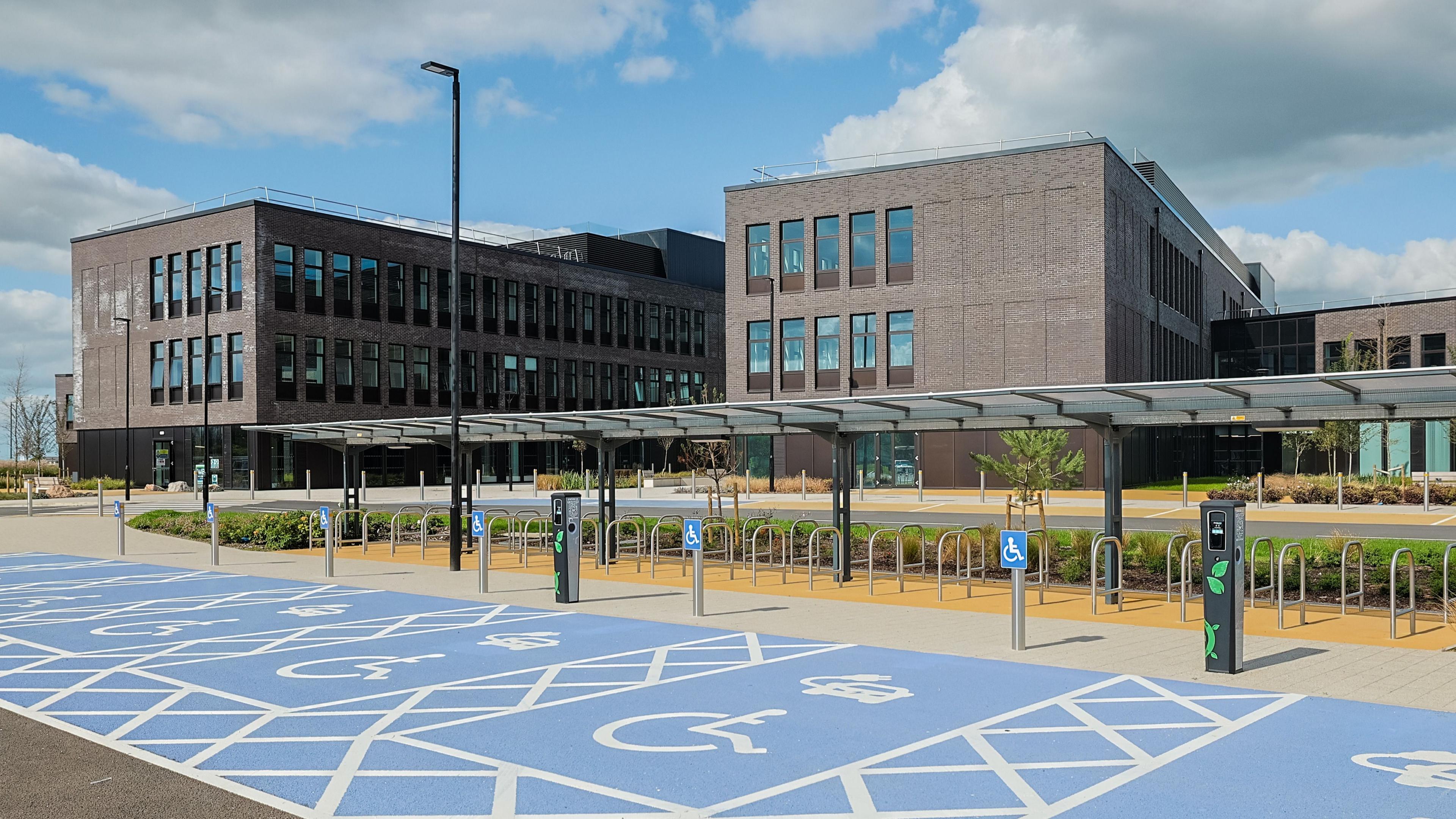 A computer-generated image of Pentre Awel, showing a car park with disabled spaces and cycle parking in front of a series of modern, brown-brick buildings.