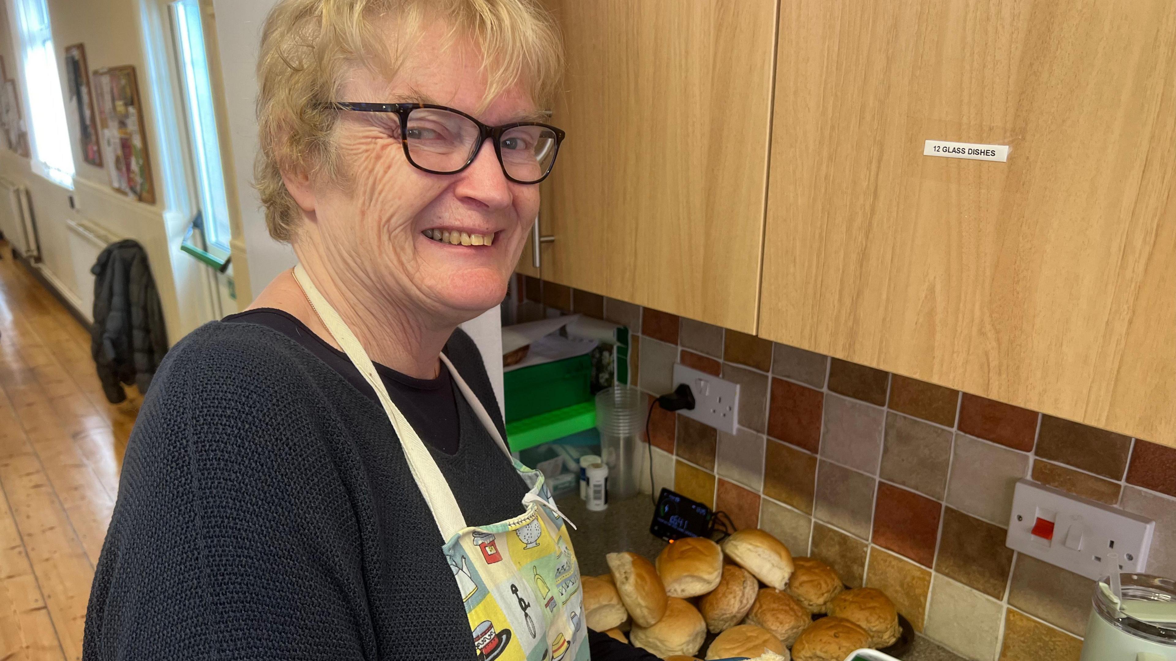 Fiona Goldsmith. She has short blonde hair and glasses. She is wearing a navy blue jumper and an apron. She is turning her head to the side to look at the camera and is smiling. A plate full of bread rolls can be seen on the counter in front of her.