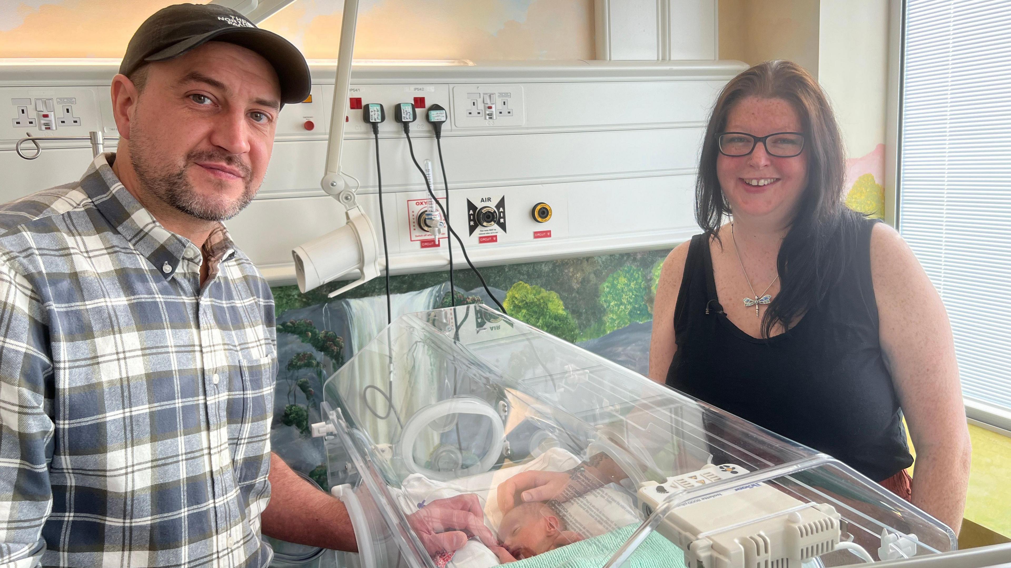 Luke and Becky standing either side of an intensive care hospital cot where baby Noah is lying. Luke has a dark goatee and is wearing a black cap and checked shirt. Becky has long dark hair and is wearing black framed glasses and a black vest with a silver necklace. Both of them have their hands placed inside the cot, touching Noah's head and hands. 