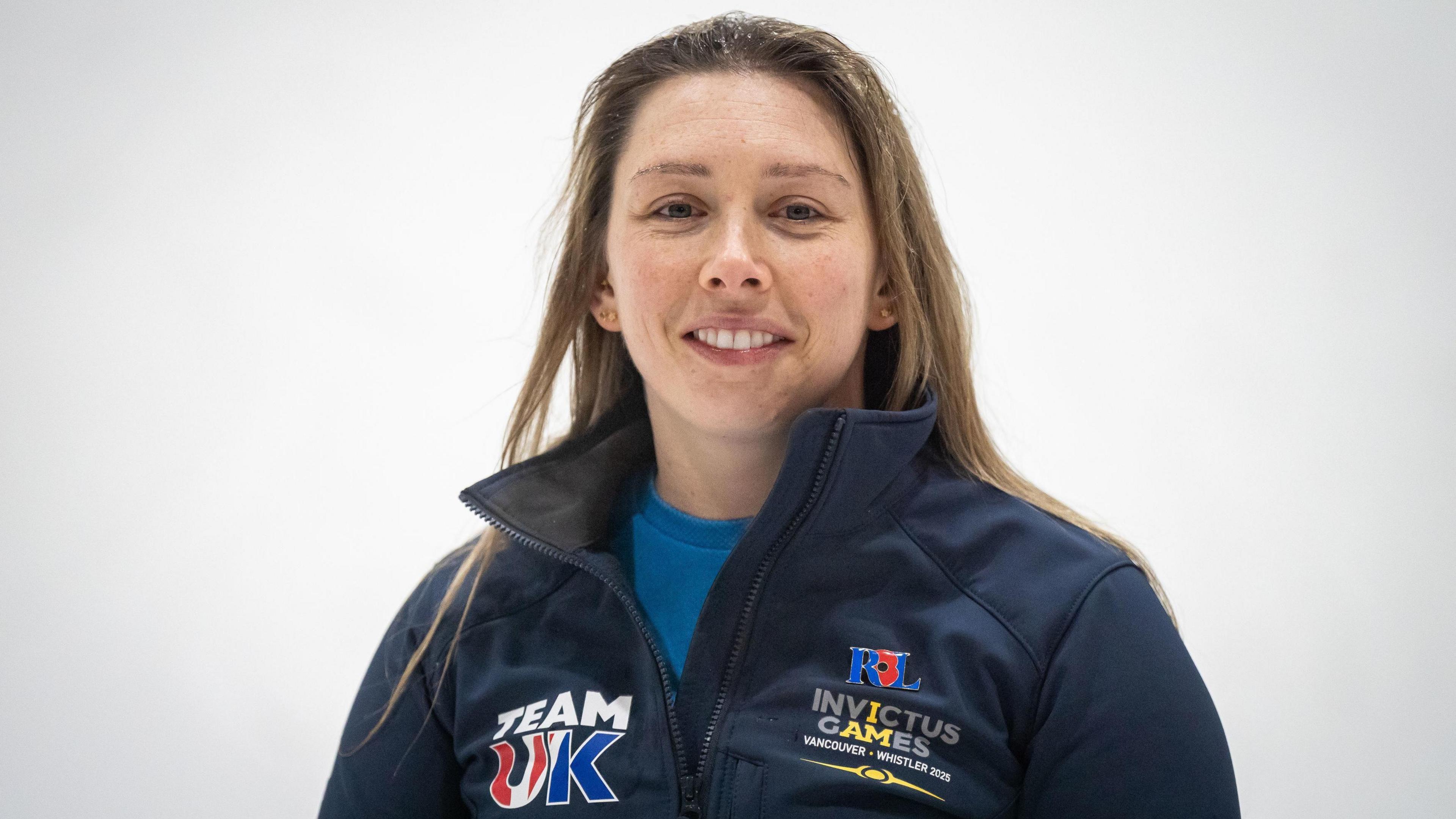 A woman with long dark blonde hair is wearing a navy blue fleece over a turquoise top. Her fleece is labelled with Team UK and Invictus Games badges.