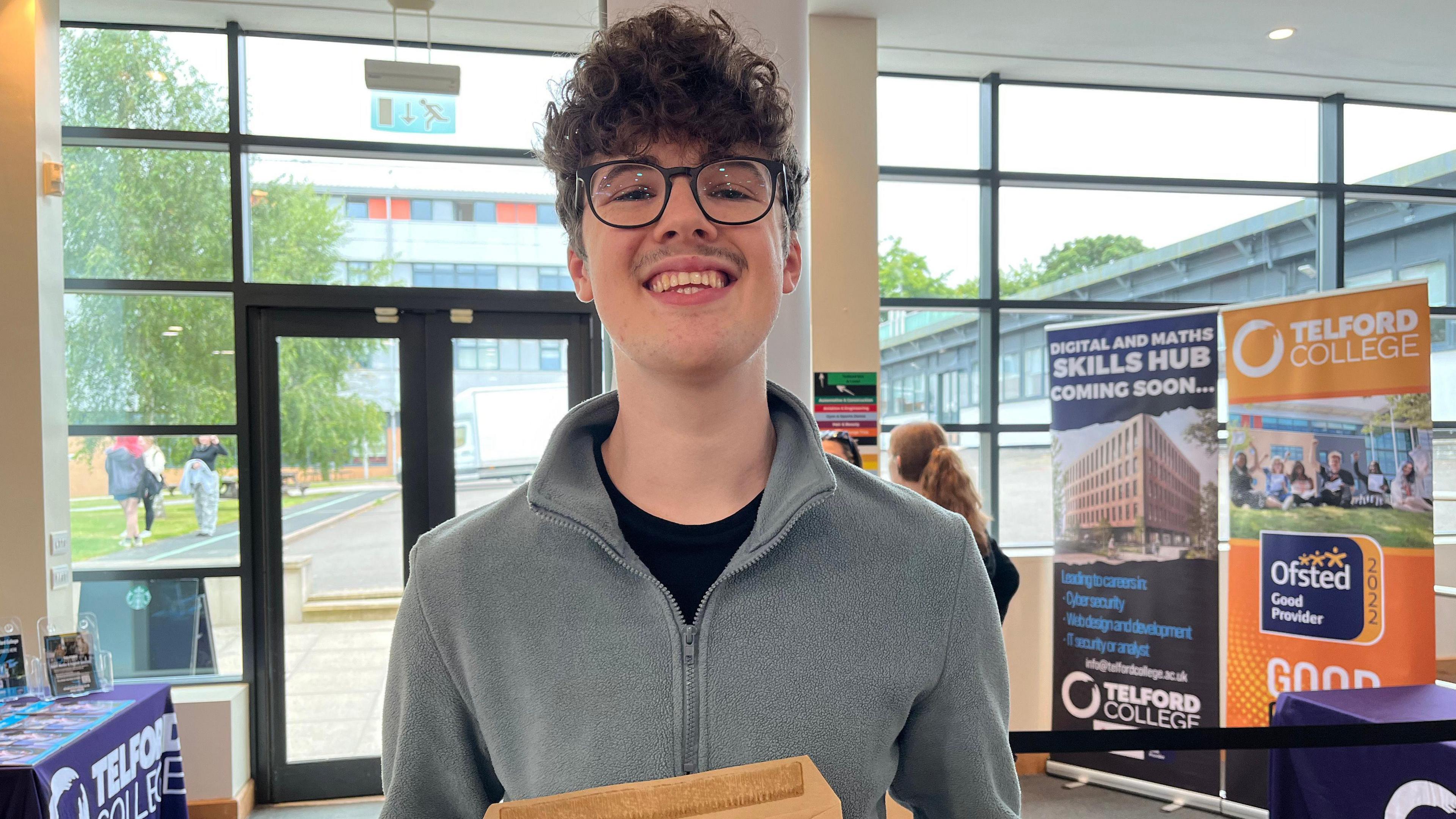 Gabriel Fay is smiling into the camera. He is wearing a grey zip fleece with a black t-shirt. He has black glasses and curly brown hair. He is holding a light brown envelope. Behind him are glass windows, and banners that read "Telford college".